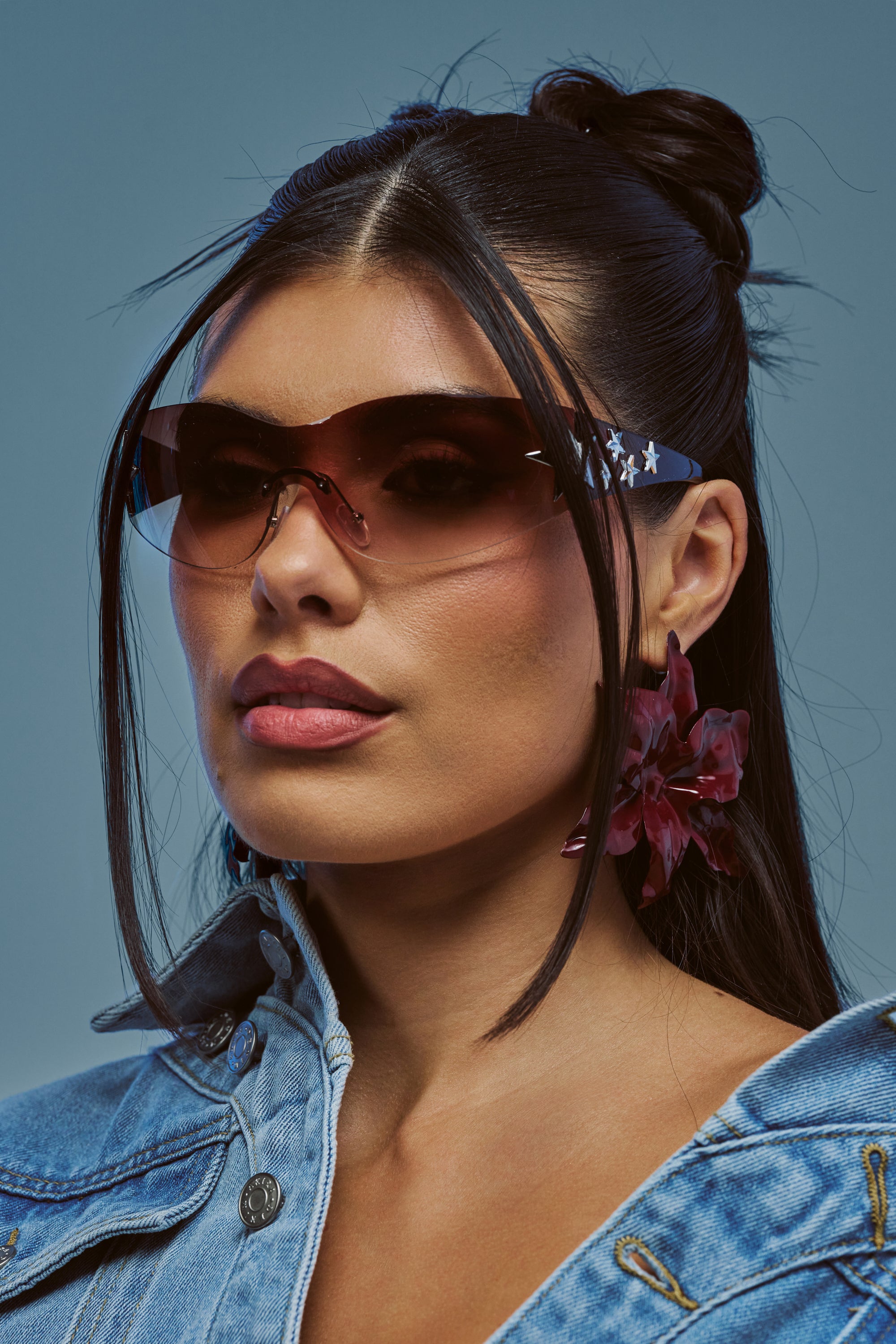 A woman with long dark hair styled half-up poses against a blue background, wearing the HOTTIE ALERT SUNGLASSES—large star-shield frames with maroon-tinted lenses—paired with bold floral earrings and a denim jacket.