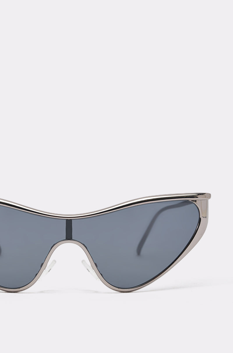 Close-up of the TO THE FUTURE SUNGLASSES showing a sleek silver-tone metal frame and dark tinted lens, set against a plain light gray background. Only part of the frame and one lens are visible. 3