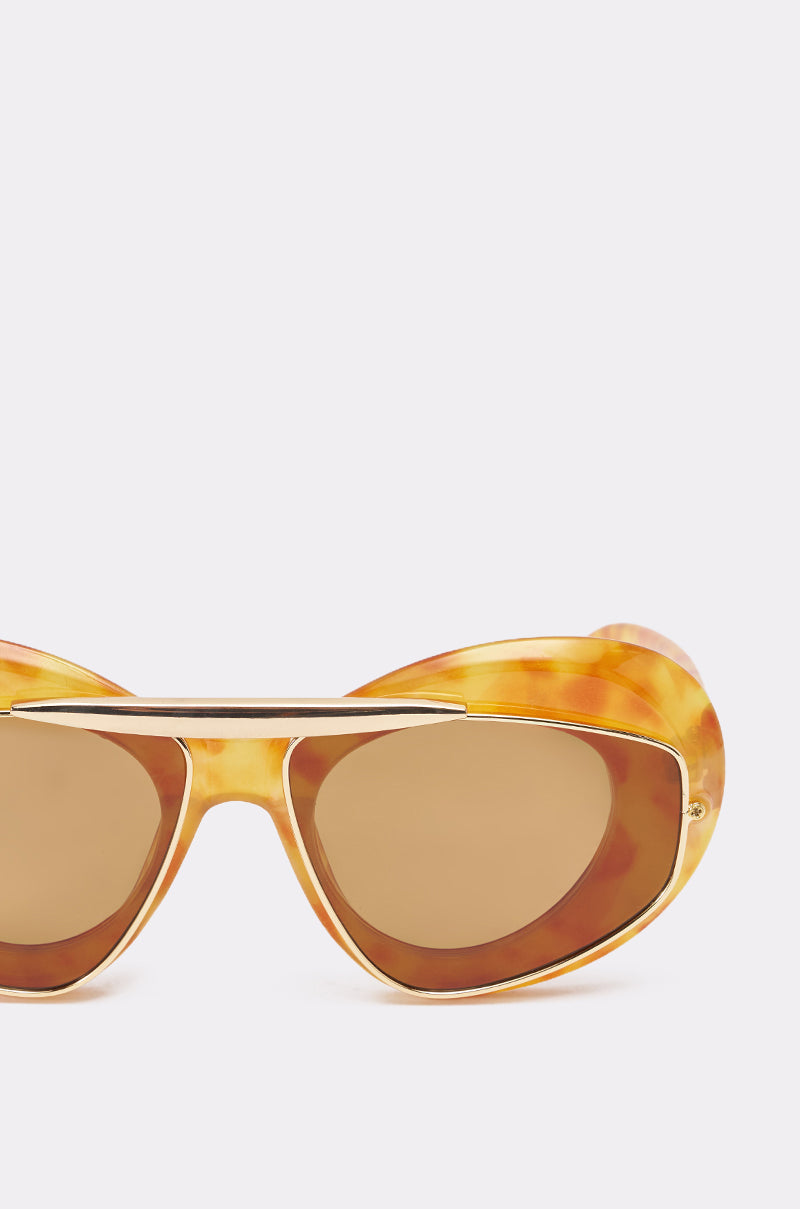 Close-up of QUITE LUX SUNGLASSES with brown-tinted lenses, oversized tortoise shell frame, and a distinctive gold-tone brow bar above the bridge, set against a plain light background. 3