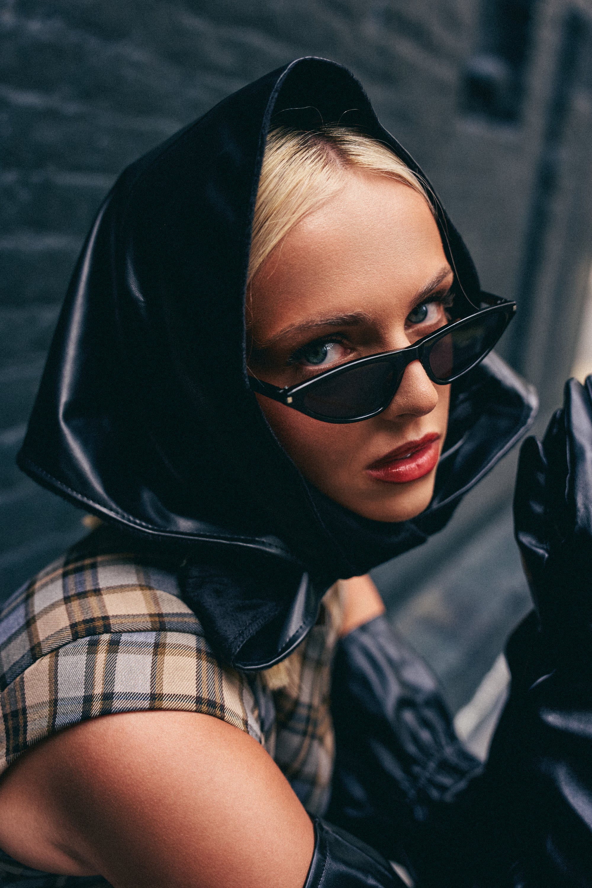 A woman wearing a black headscarf, chic summer accessories including the RUNWAY READY SUNGLASSES, a plaid top, and black gloves looks intensely at the camera against a blurred urban background.