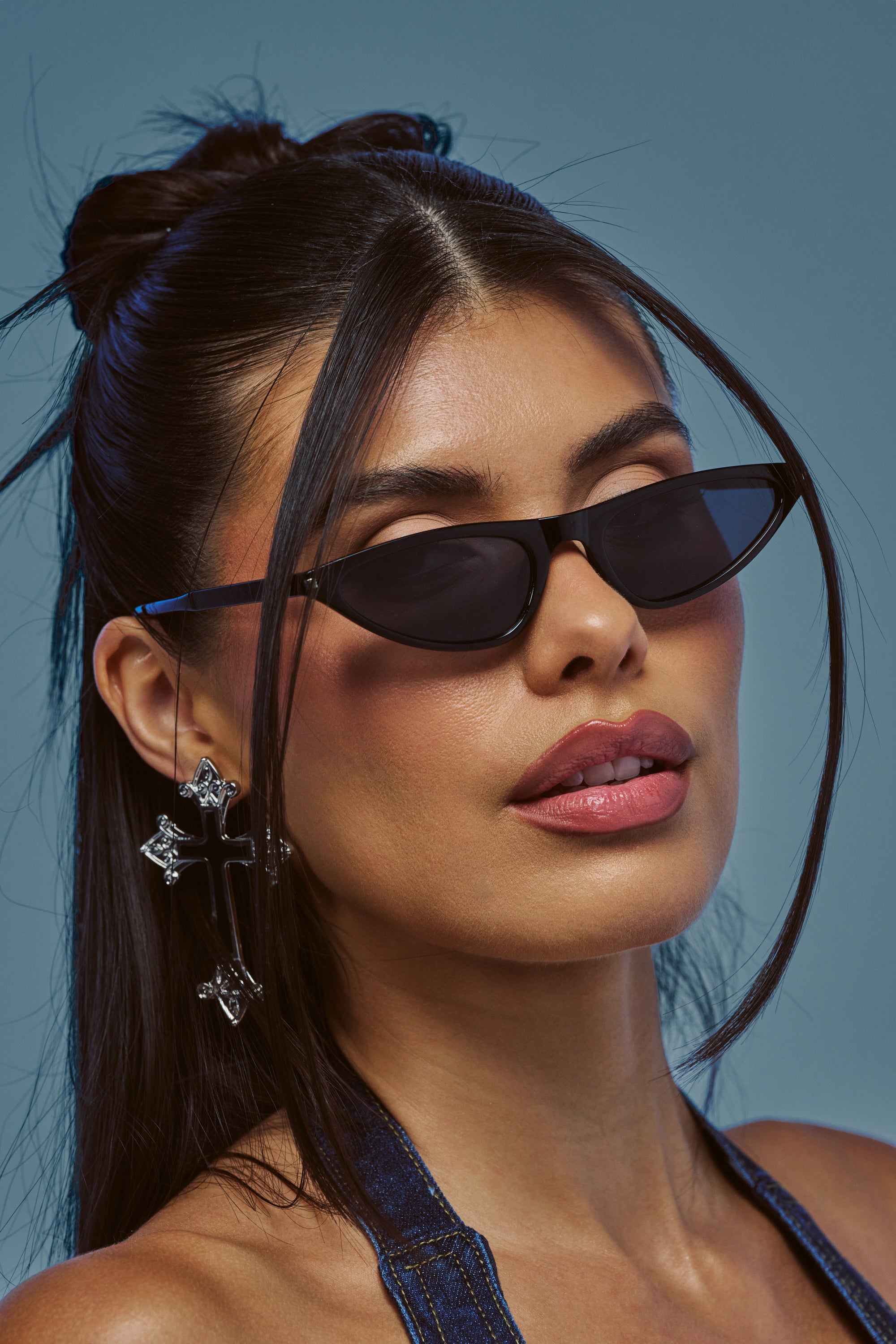 Woman with long dark hair in a half-up style, wearing RUNWAY READY SUNGLASSES, cross-shaped earrings, and a halter denim top, posing against a blue background.