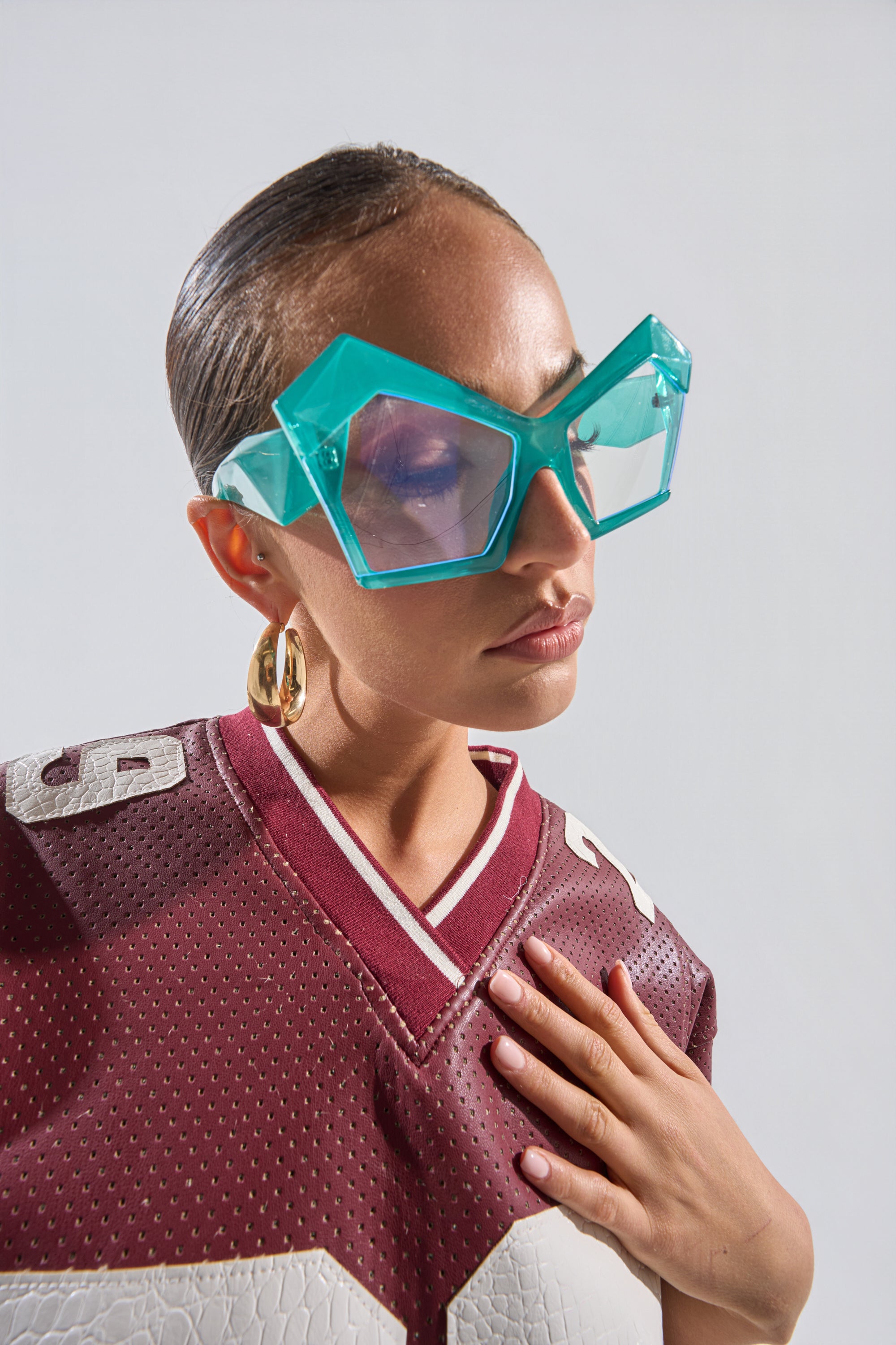 A woman wearing KYLEIGH GLASSES with oversized square geometric frames, a maroon football jersey, large gold hoops, and slicked-back hair poses with her hand on her chest against a plain background. 0