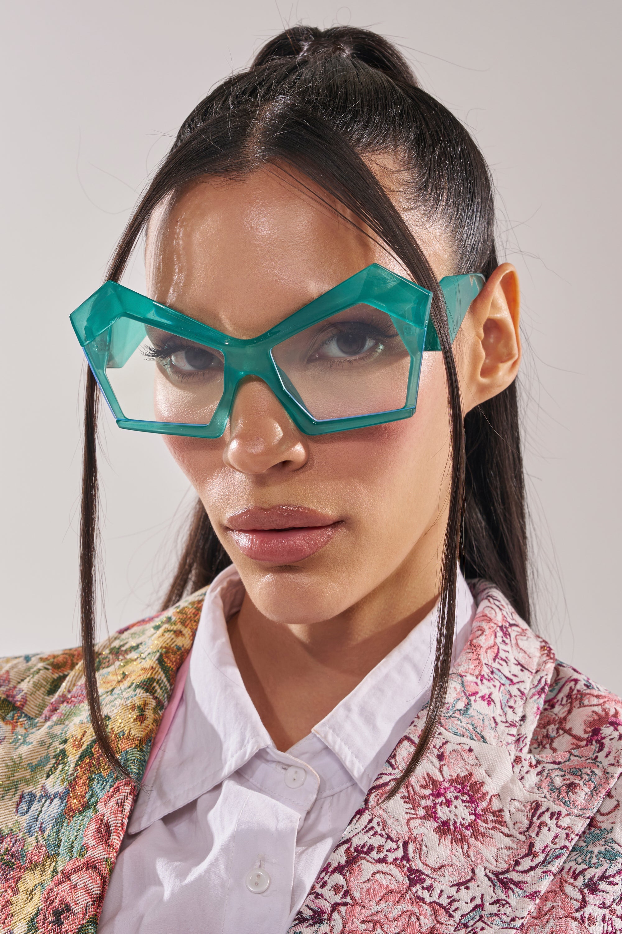A woman with long dark hair in a high ponytail wears oversized, angular KYLEIGH GLASSES and a floral jacket over a white collared shirt, posing against a plain light background. 2