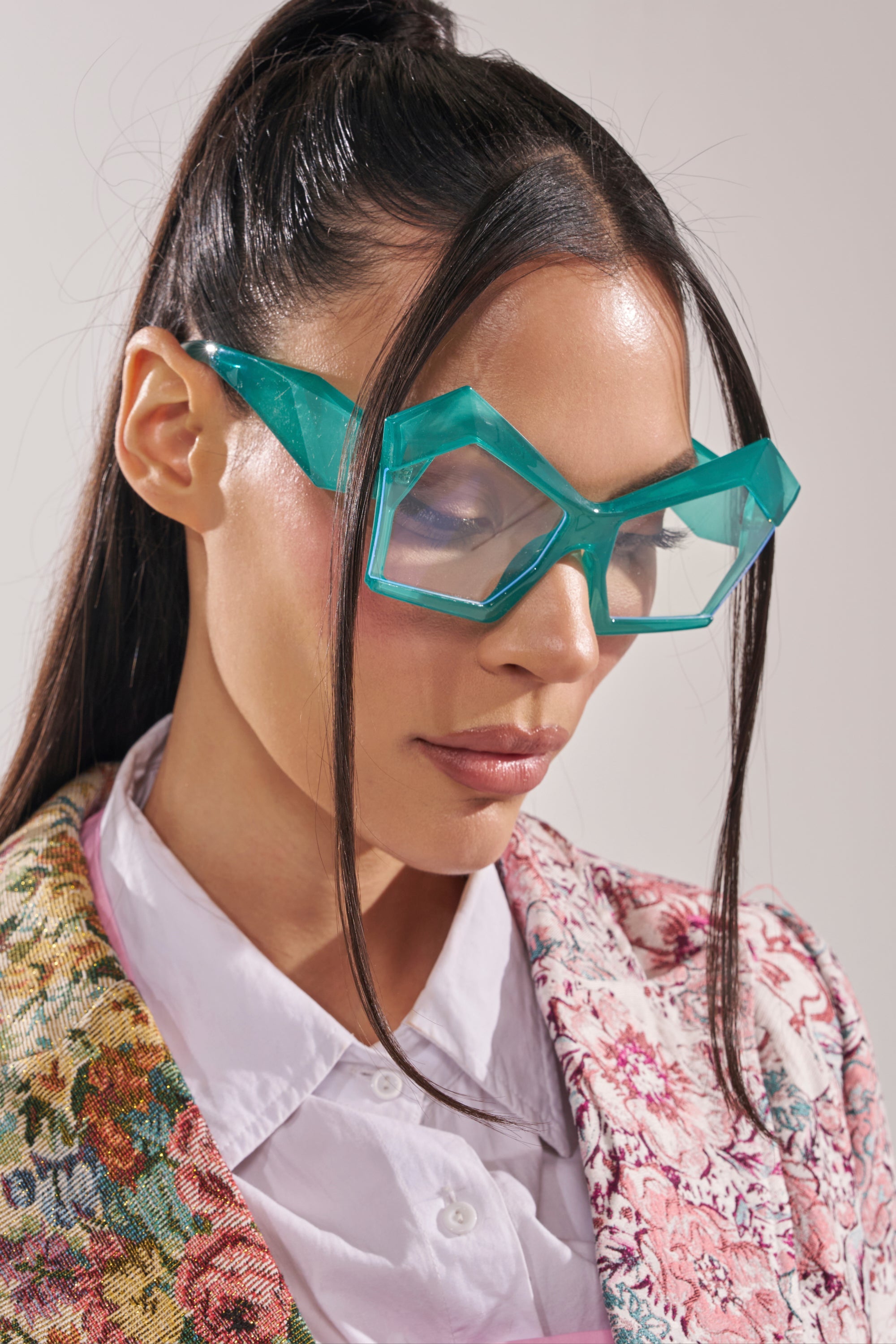 A woman with long dark hair wears bold, oversized KYLEIGH GLASSES in turquoise and a floral textured jacket over a white collared shirt, looking down with a neutral expression. 3