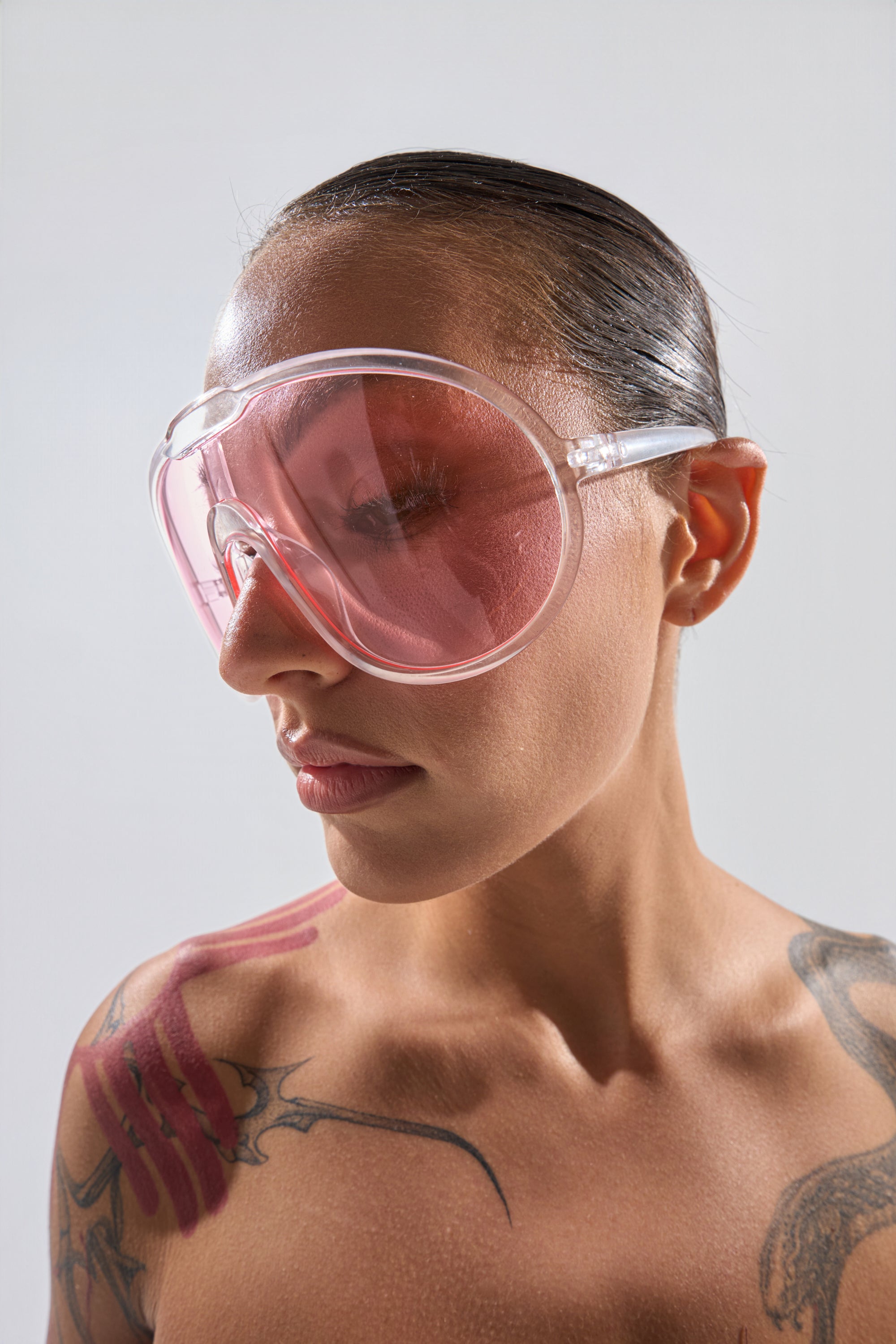 A woman with slicked-back hair wears PINK VISION SUNGLASSES, highlighting her shoulder and collarbone tattoos as she poses against a plain, light background. 0