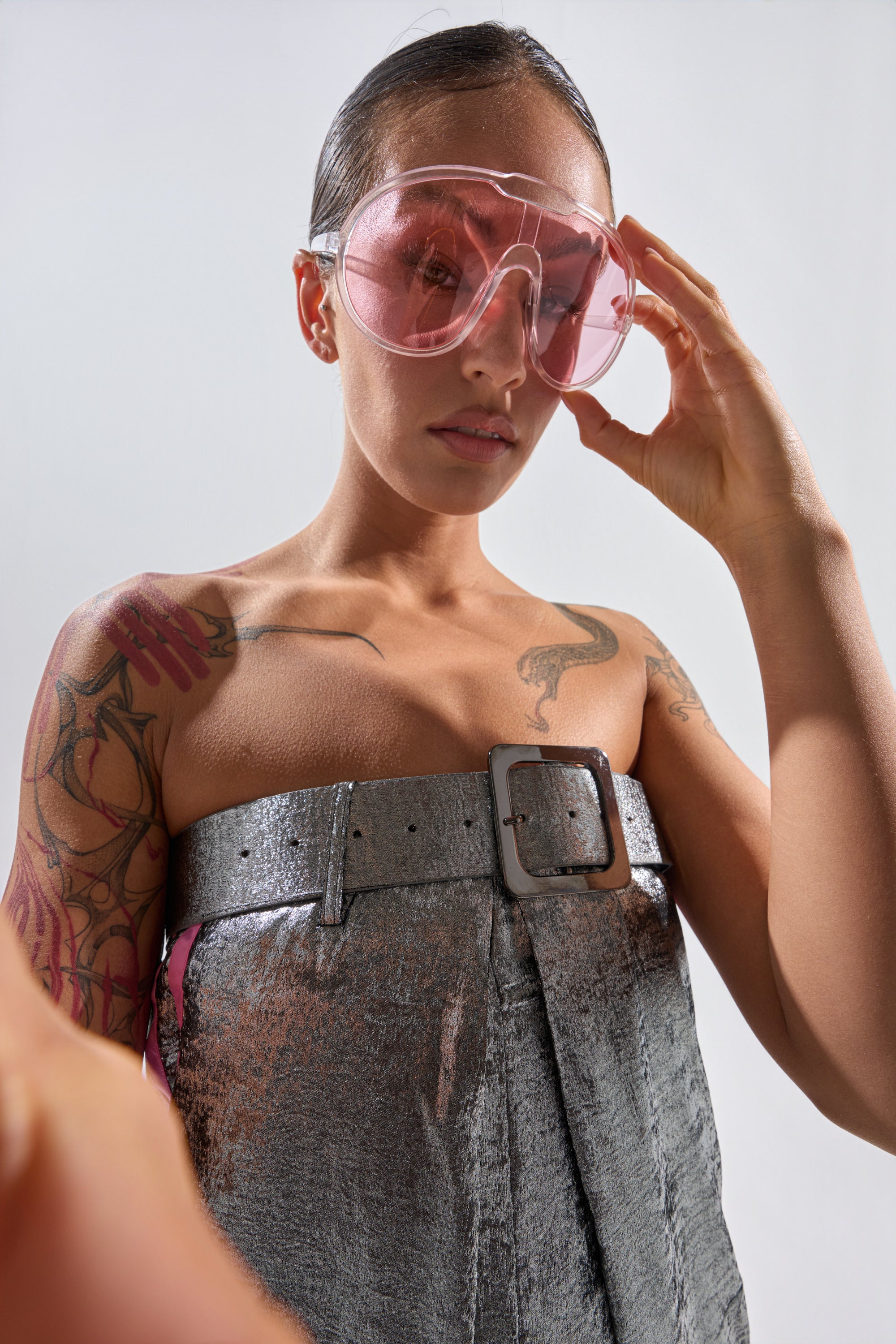 A tattooed person wears PINK VISION SUNGLASSES and a metallic belted strapless top, looking down at the camera while holding the sunglasses with one hand against a plain light background. 1