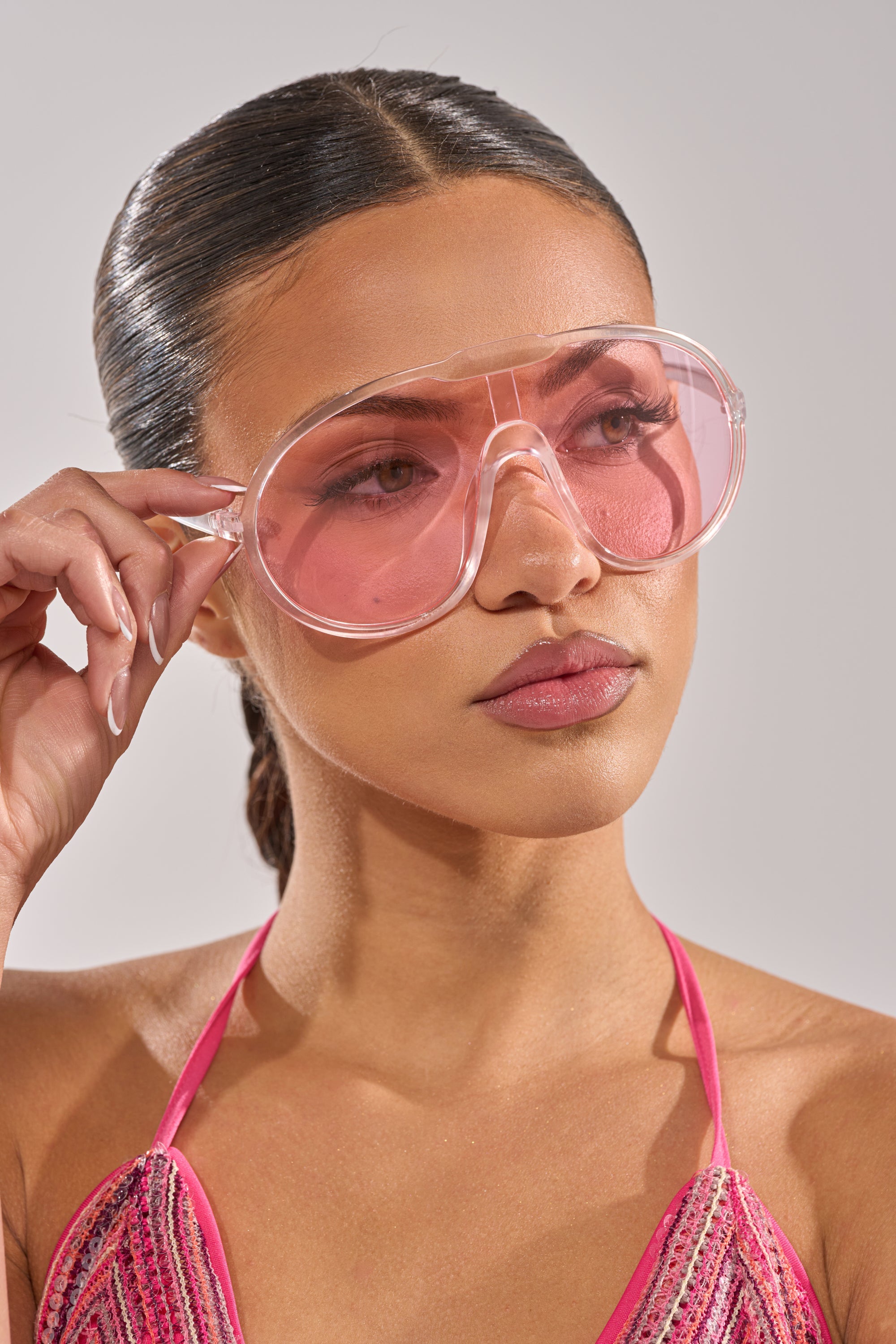 A woman with sleek, dark hair in a low ponytail wears PINK VISION SUNGLASSES—large, translucent pink frames—paired with a pink halter top. She looks to the side, holding the sunglasses with one hand against a plain background. 3