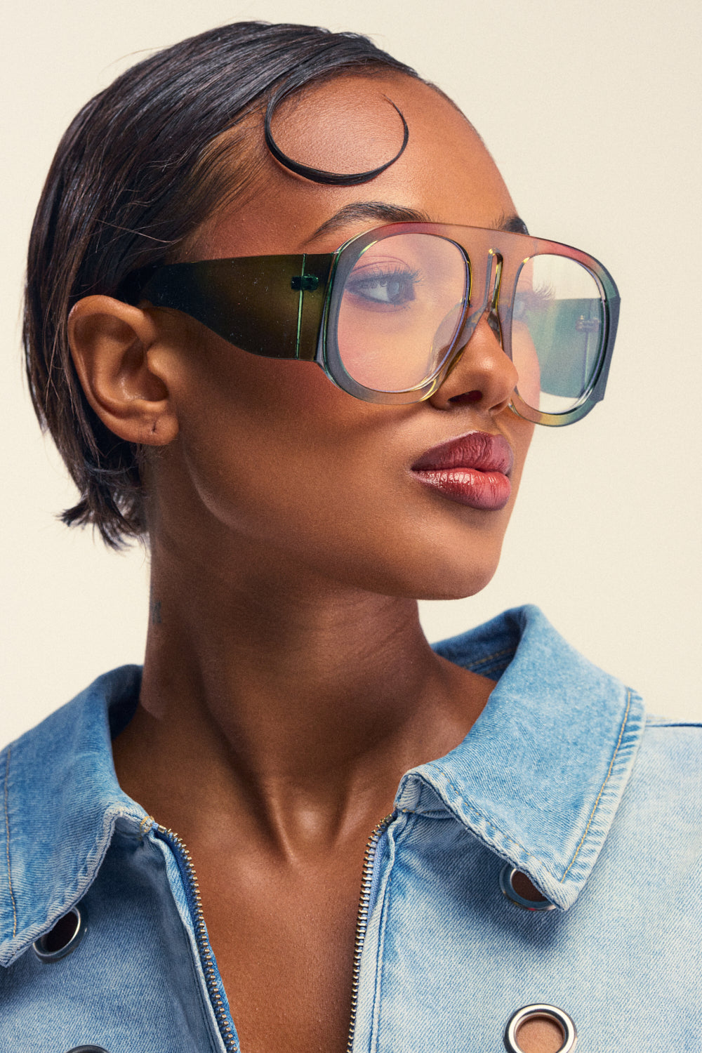 A close-up portrait of a woman with a sleek hairstyle, wearing the bold and fashionable IT GIRL GLASSES in an oversized green frame and a denim jacket, looking to the side against a neutral, cream-colored background. The statement IT GIRL GLASSES elevate her modern look.