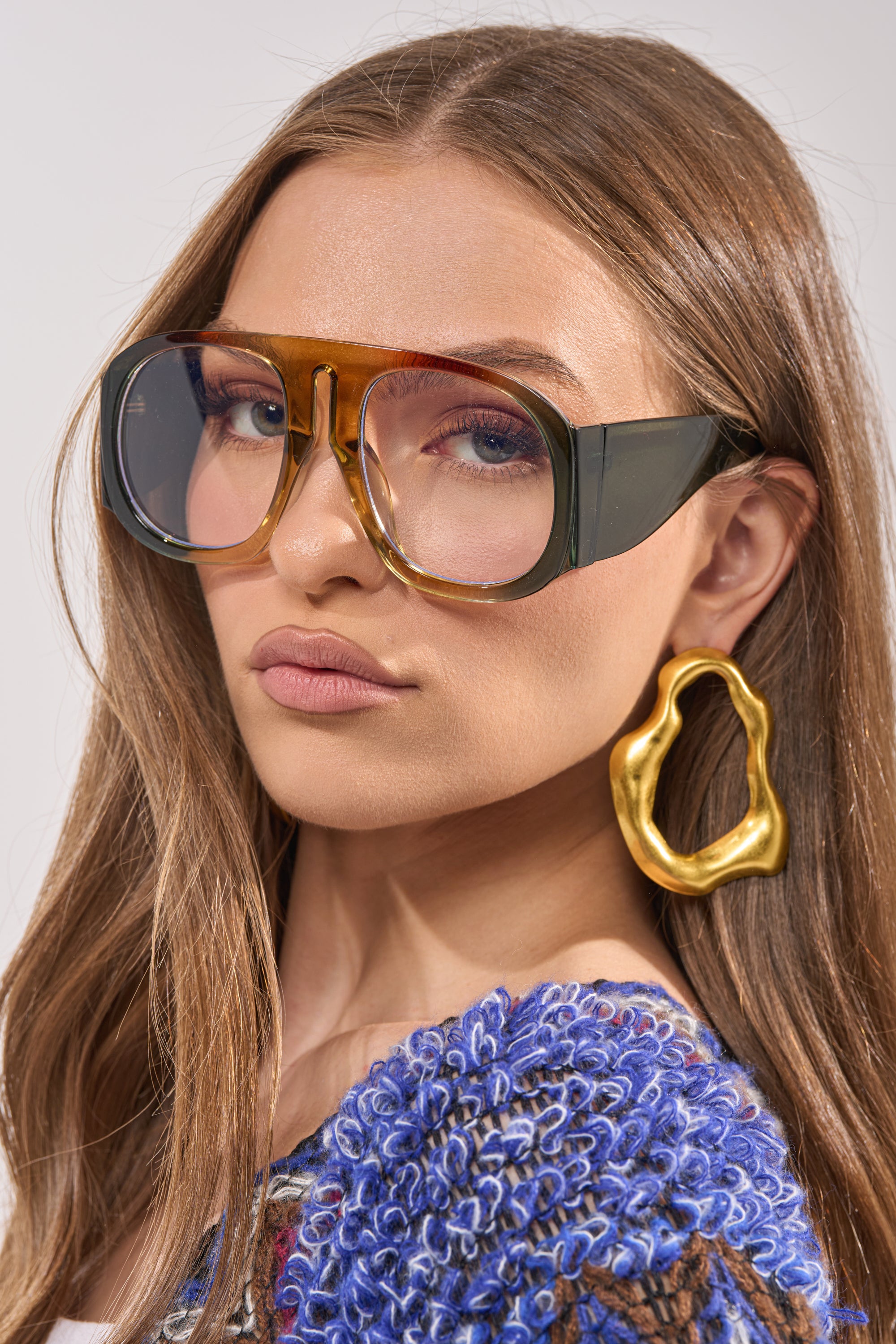 A woman with long brown hair wears IT GIRL GLASSES—oversized, two-tone statement eyewear—paired with large gold abstract earrings and a textured blue-patterned top, looking confidently at the camera against a neutral background. 2