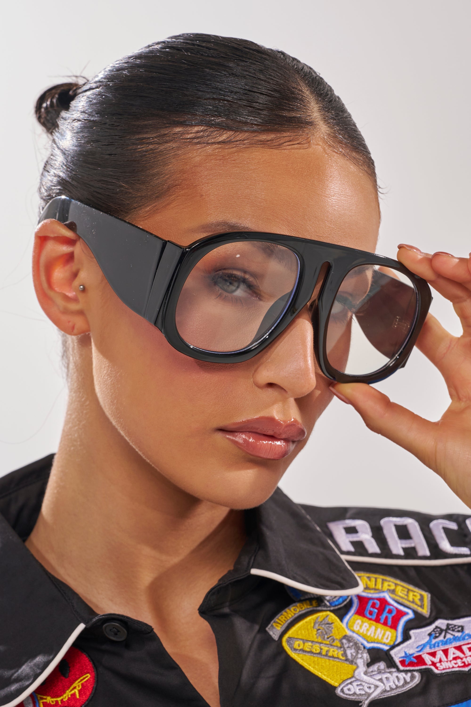 A woman with slicked-back hair confidently adjusts her READ ME GLASSES—black oversized sunglasses with a glossy frame—while wearing a black shirt adorned with colorful racing patches and looking at the camera. 1