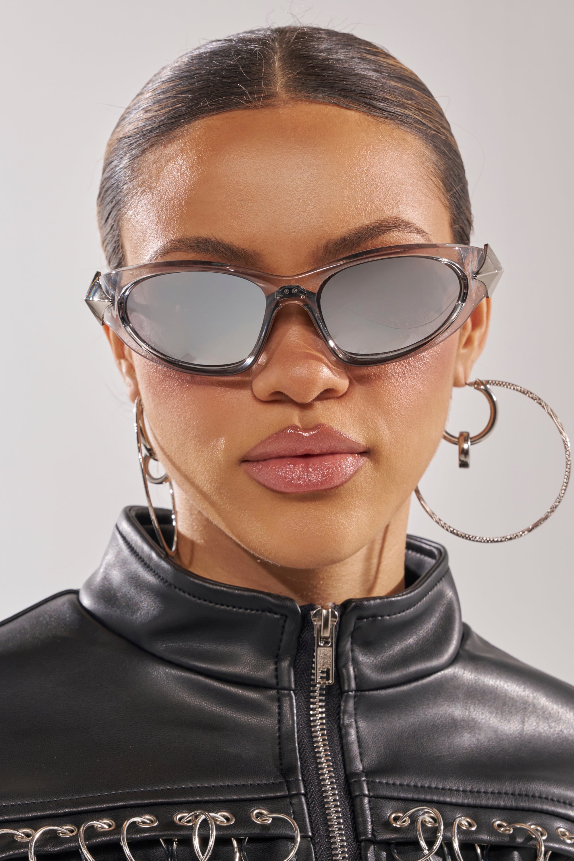 A woman wears STARSTRUCK SUNGLASSES with mirrored lenses, silver hoop earrings, and a black faux leather zip-up jacket, looking straight at the camera against a light background. 1