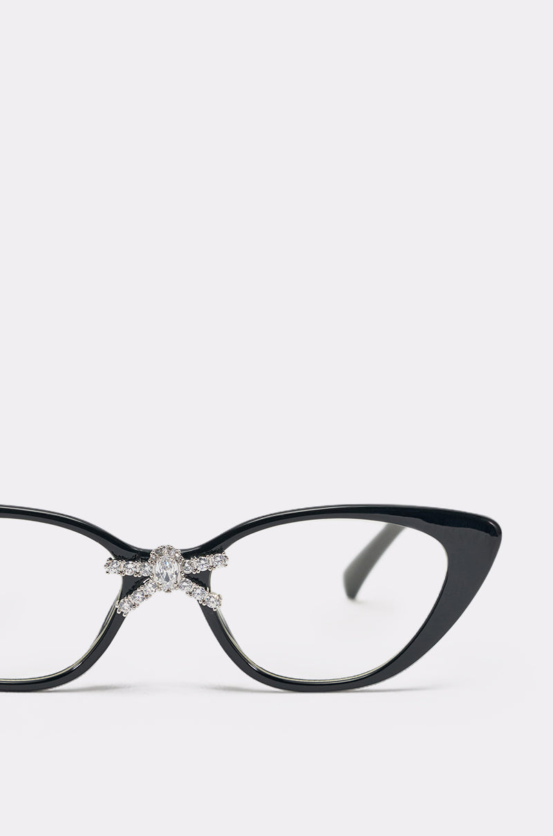 A close-up of FEMME GLASSES, featuring black cat-eye frames with rhinestone accents and a sparkling bow embellishment above the nose bridge, set against a plain light background. 4