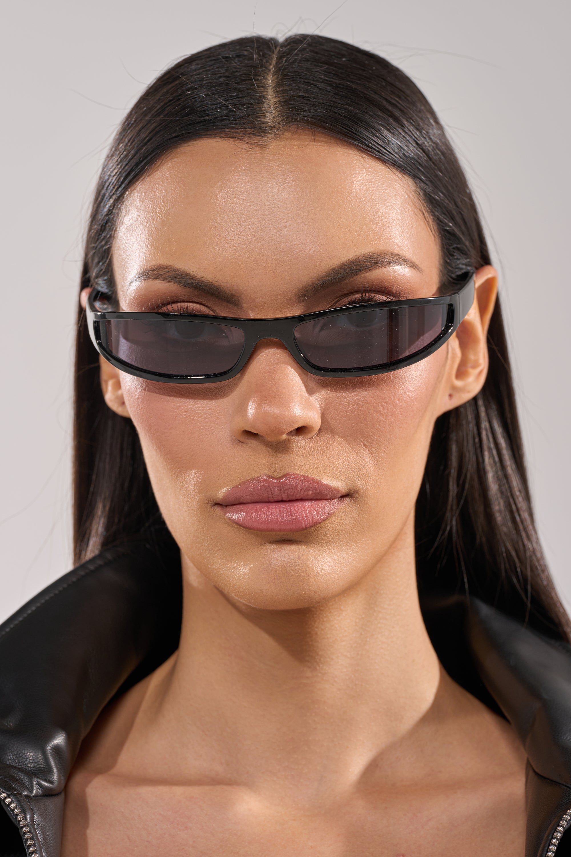 A woman with straight dark hair wears IN THE FUTURE SUNGLASSES, a black leather jacket with the collar turned up, and neutral makeup, looking directly at the camera against a plain background. 1