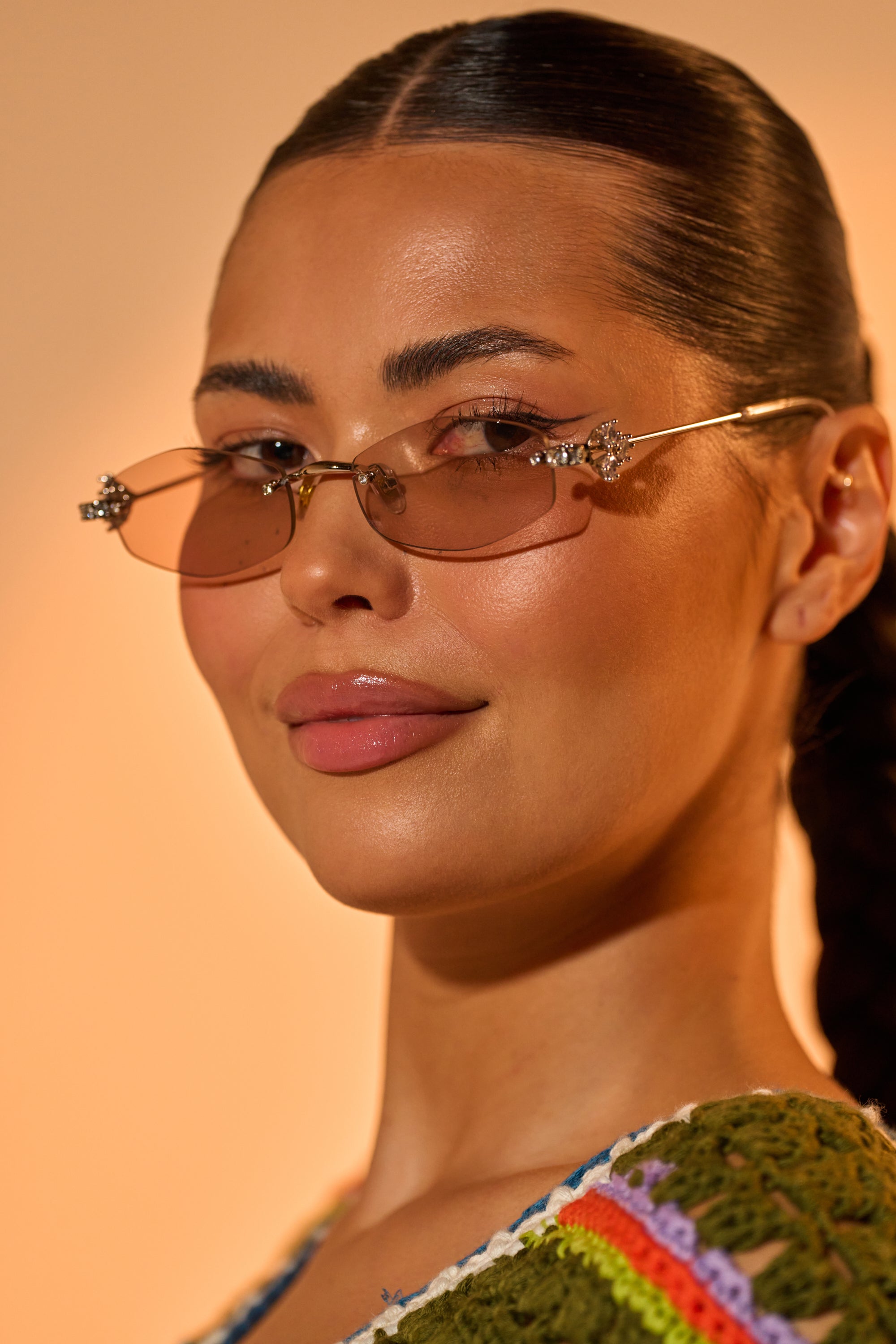 A woman with slicked-back dark hair models TRENDSTER GLASSES—narrow Y2K ovals with brown tinted lenses and jeweled accents. She wears a glossy lip and green crochet top with colorful stripes, posed against a warm beige background. 2