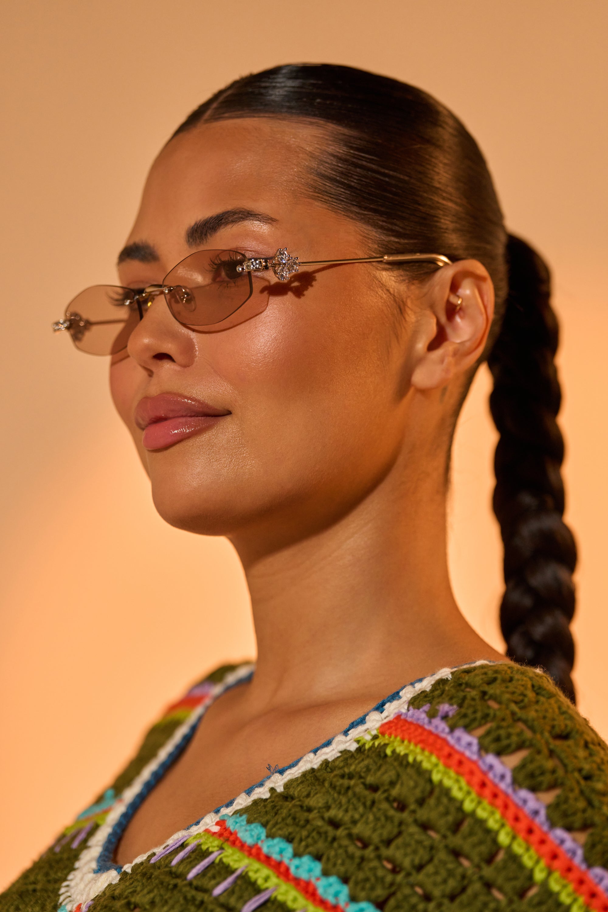 A woman with long dark braided hair wears TRENDSTER GLASSES with brown tinted lenses and a green multicolored crocheted top, facing sideways confidently against a warm neutral background. 3