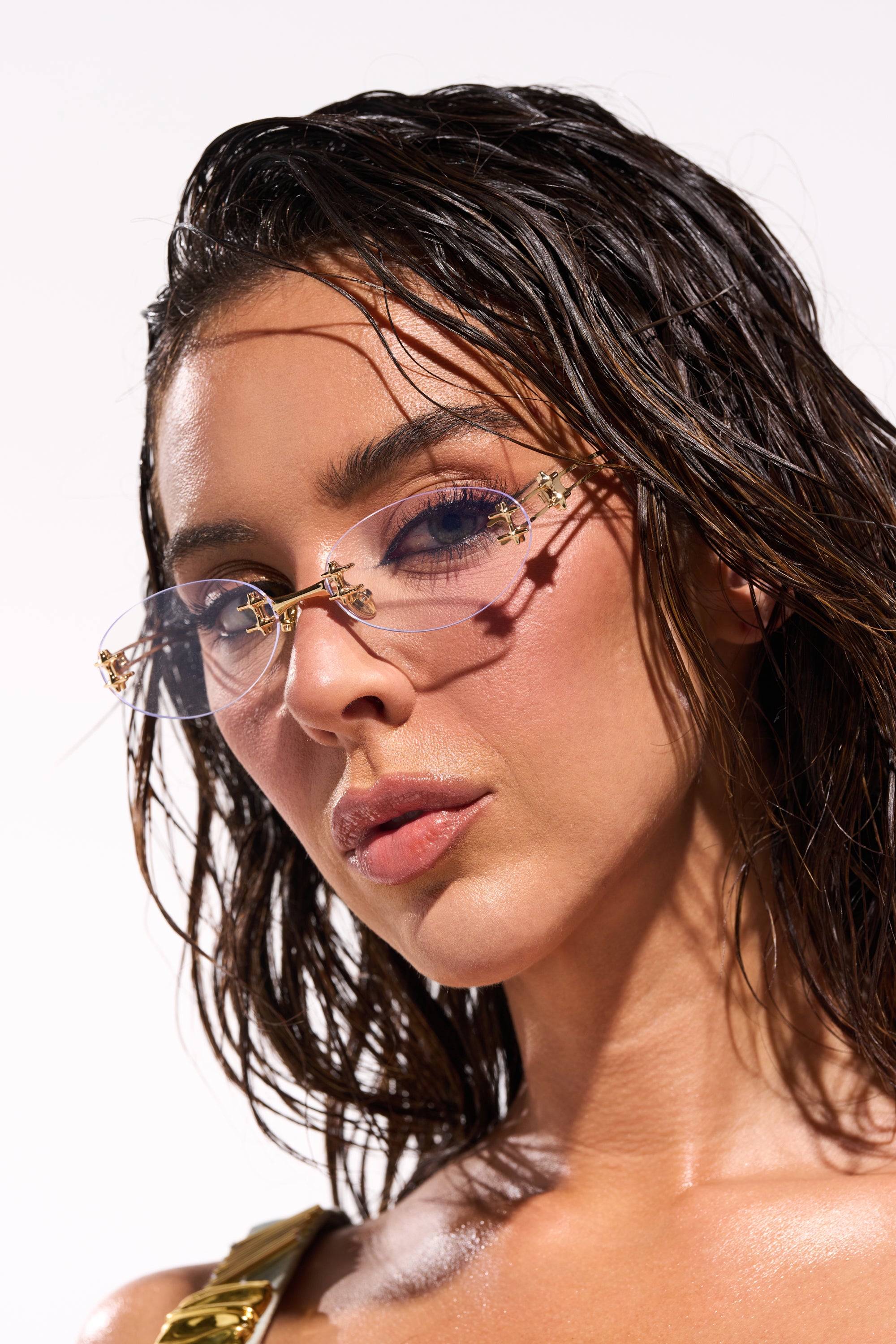 A woman with wet, wavy dark hair wears TREND DRIVEN GLASSES—minimalist sunglasses featuring a gold-tone metal frame and decorative details. Her dewy skin and confident expression stand out against a plain light background. 0