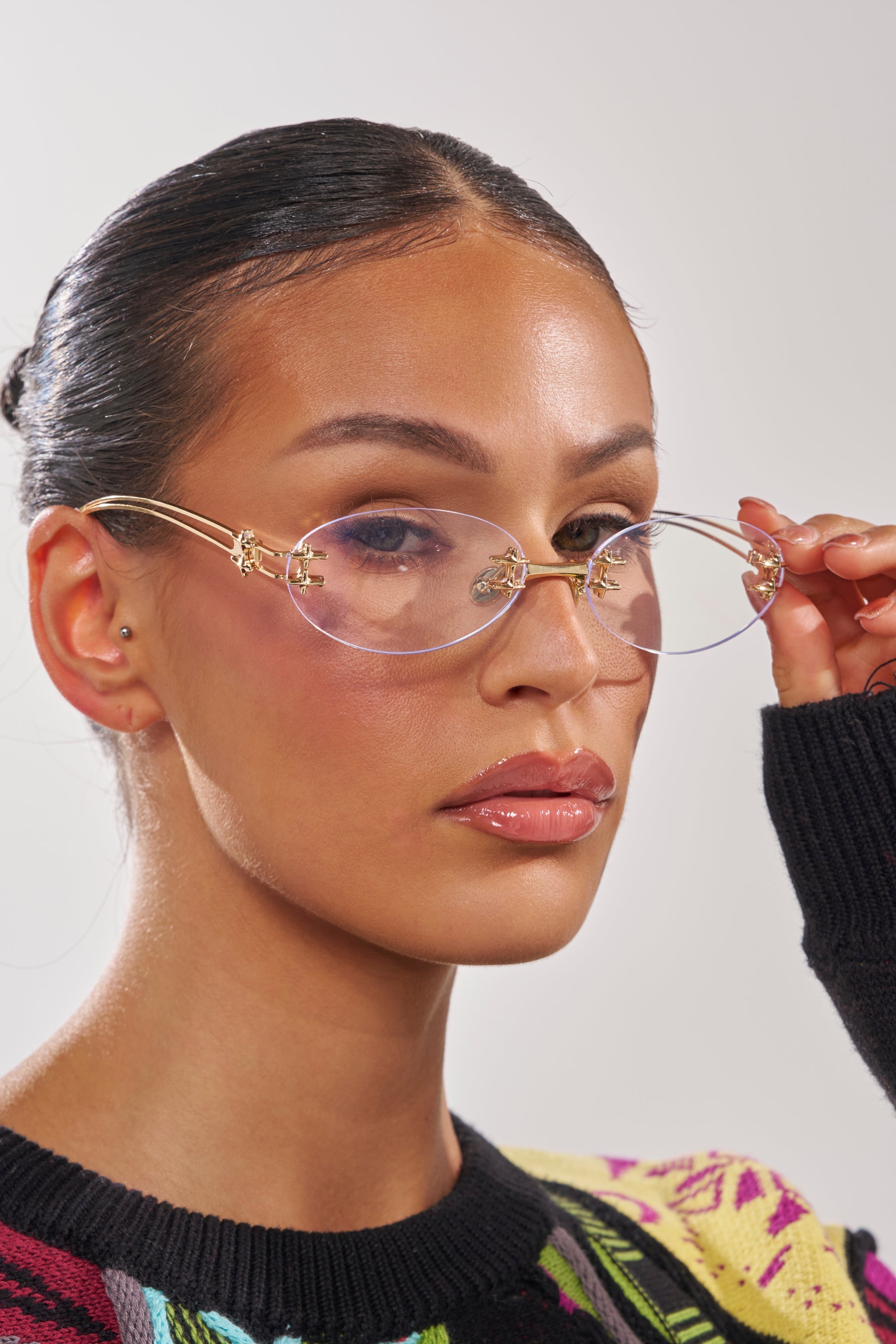 A confident person adjusts their TREND DRIVEN GLASSES with one hand, showcasing smooth pulled-back hair, glossy lips, and a black sweater with colorful knit accents against a plain light background. 3