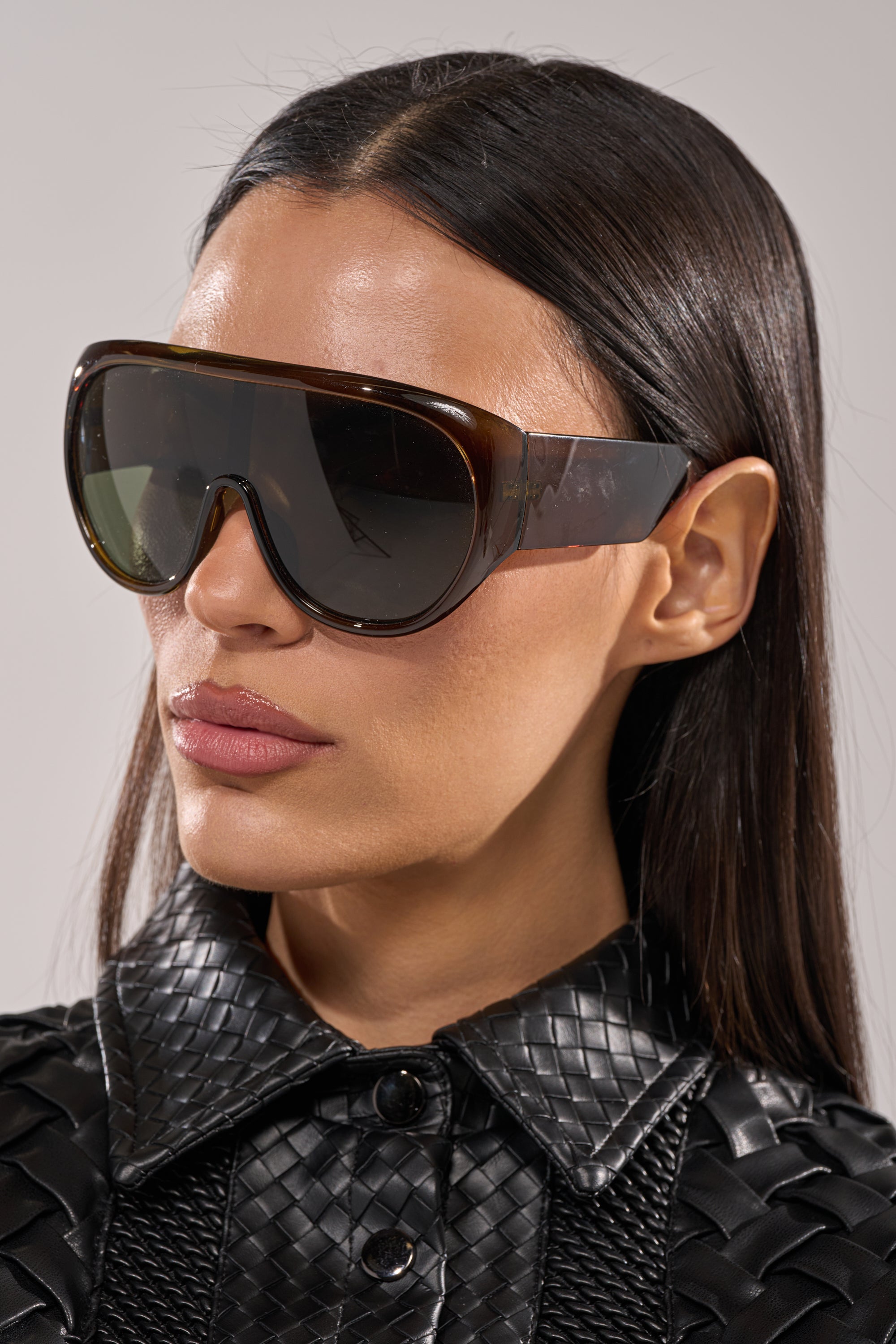 A woman with straight dark hair wears oversized TOMBOY SUNGLASSES and a black, textured faux-leather jacket, facing slightly to the side with a neutral expression against a plain background. 2
