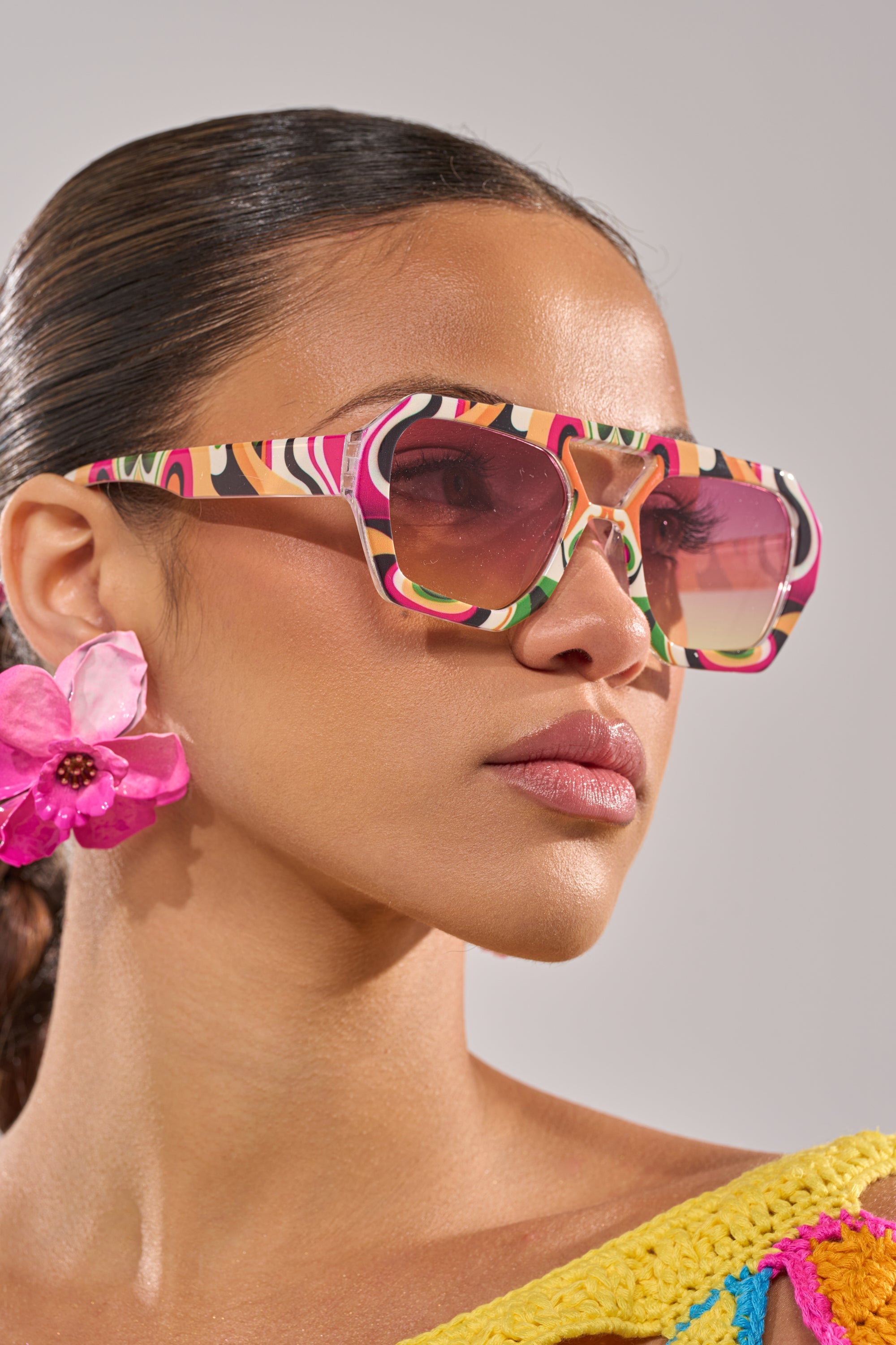 A woman with sleek, pulled-back hair wears FEEL THE BEAT SUNGLASSES—oversized and vibrant—and a bright pink flower earring, accenting her smooth complexion and colorful multicolored outfit. 2