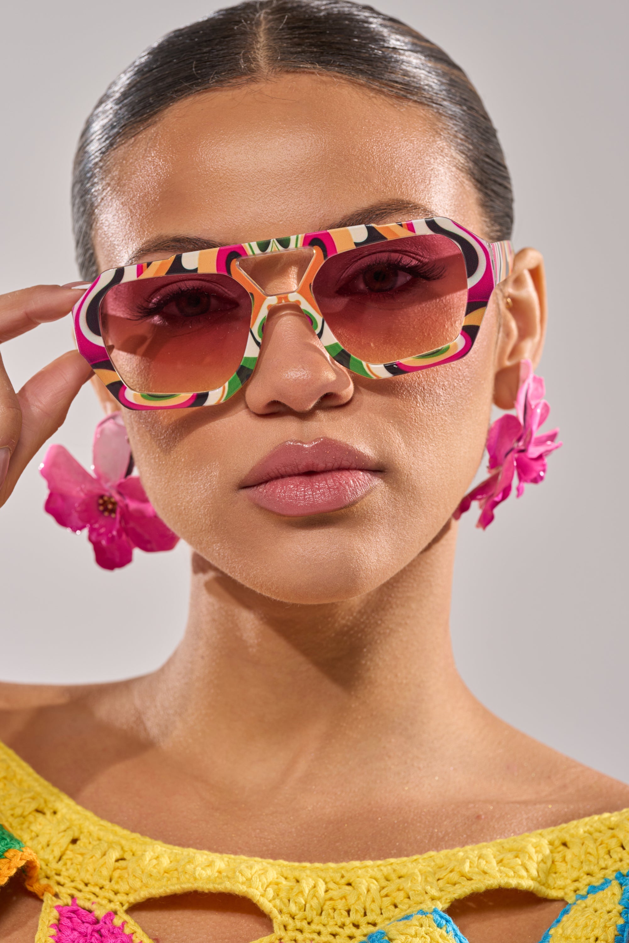 A woman in slicked-back hair and a yellow crocheted top confidently poses in FEEL THE BEAT SUNGLASSES, multicolor square frames, paired with bold pink flower earrings against a softly lit plain background. 3