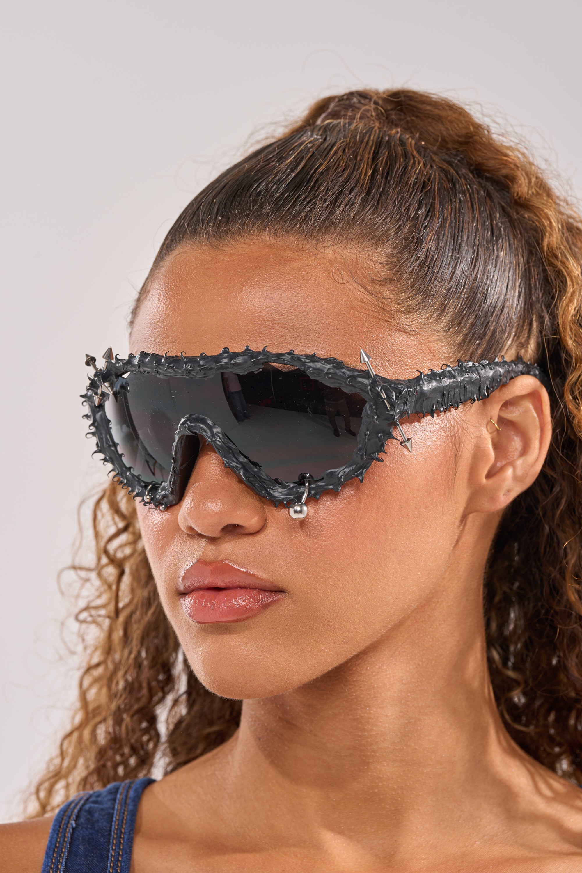 A person with curly brown hair wears DANGER SPIKES SUNGLASSES featuring large black spikes and a silver teardrop charm. They have a neutral expression and are dressed in a blue top. 1