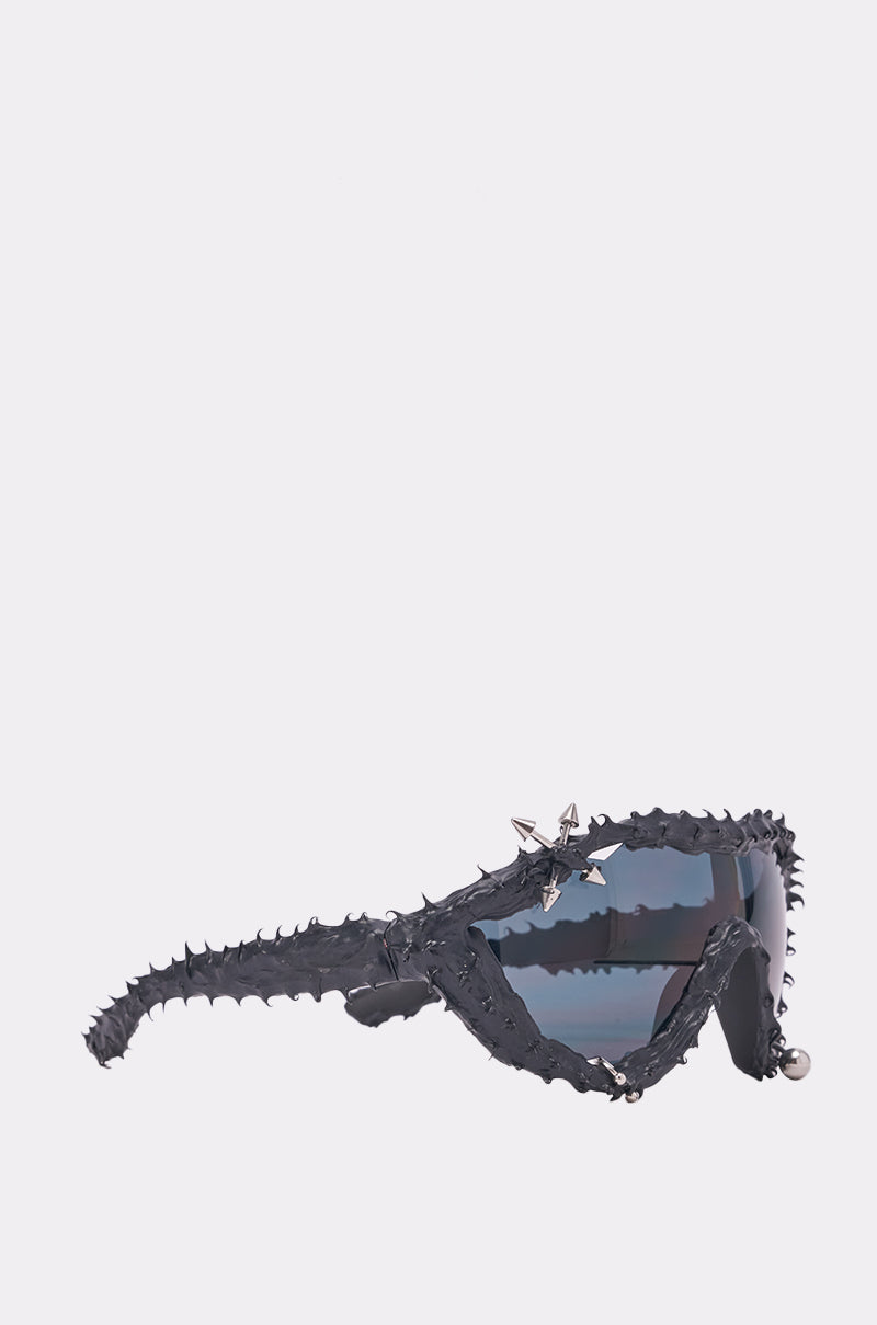 The DANGER SPIKES SUNGLASSES feature thick, angular dark frames adorned with thorn-like spikes and a small metallic star accent near the temple for a bold, futuristic look. 3