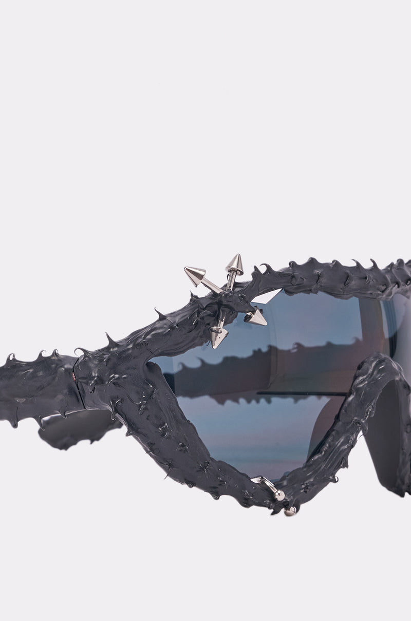 Close-up of the DANGER SPIKES SUNGLASSES featuring black, thorn-like frames with silver spike accents above tinted blue lenses, set against a plain light background. 4