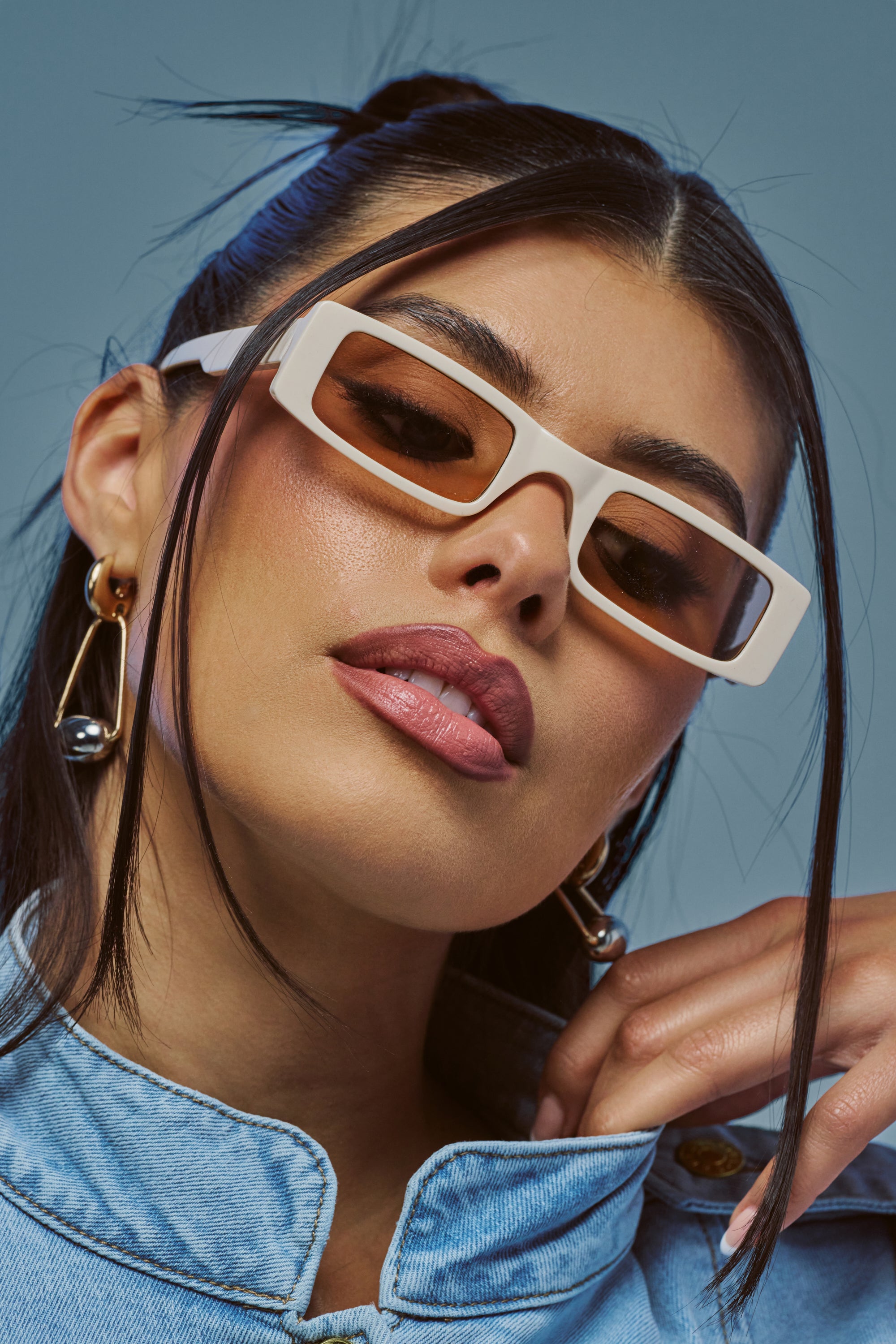 A woman poses confidently against a blue background, wearing the SUMMER LOVIN’ SUNGLASSES—chic, nude frames with rectangular tinted lenses—paired with a denim jacket. Her shiny earrings and dark hair, styled back with soft strands framing her face, complete the look.