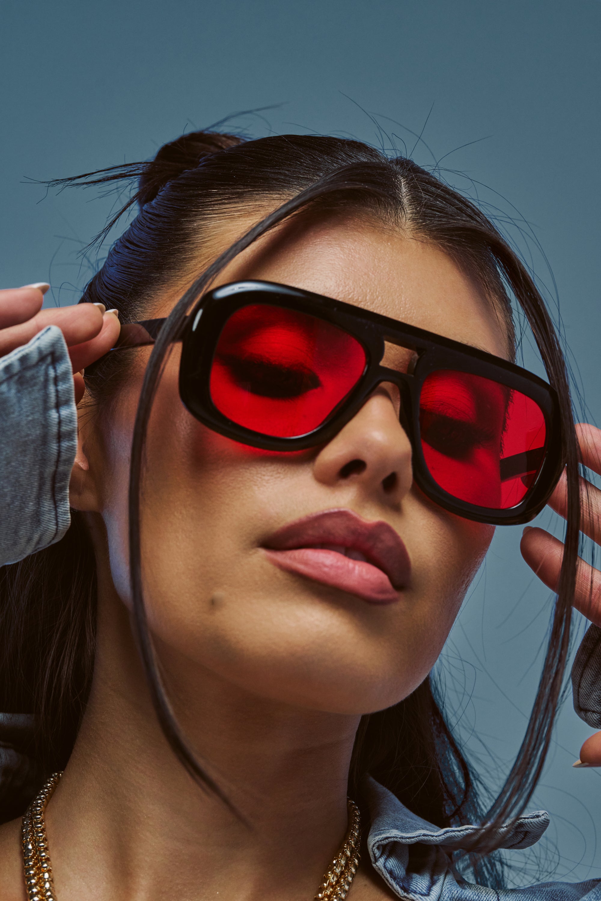 A woman wearing bold summer style showcases the RED AND HOT SHIELD SUNGLASSES—red-tinted, oversized frames—paired with a denim jacket. Her dark hair is styled back, a few loose strands framing her face, and she sports neutral makeup with a gold chain necklace as she looks confidently at the camera.
