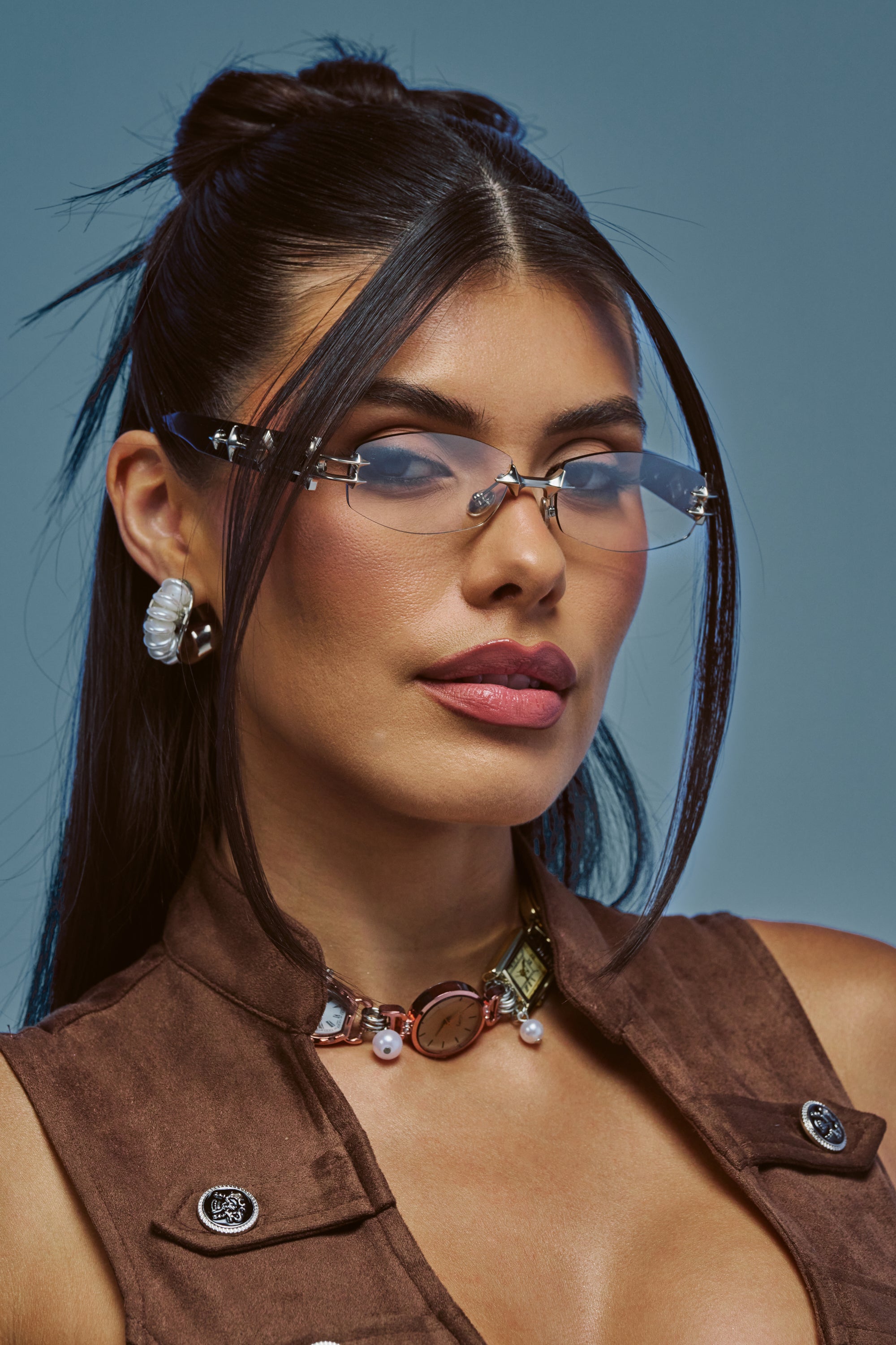 Woman with long dark hair styled half-up, wearing CHROME WHISPER FRAMES—clear glasses reminiscent of tinted narrow lens sunglasses—paired with a brown vest, statement necklace, and pearl earring, posing against a blue background.