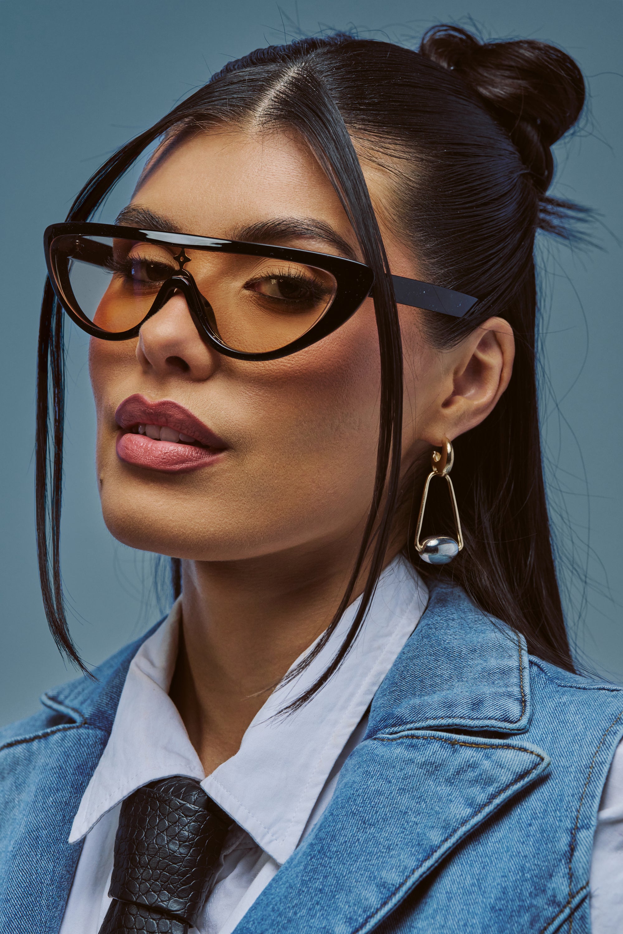 Sporting the CYBER GLIDE SUNGLASSES, a woman with straight dark hair styled in a top knot pairs her fashion-forward shades with gold earrings, a white shirt, black textured tie, and blue vest. She gazes confidently past the camera against a neutral background.