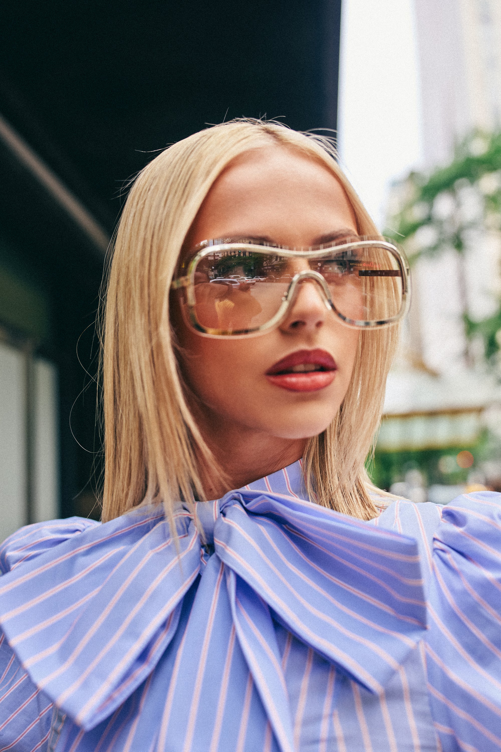 Wearing the SHEER DIVA FRAMES, a woman with straight blonde hair styles a blue blouse featuring white stripes and an oversized bow as she poses outdoors in a city setting.