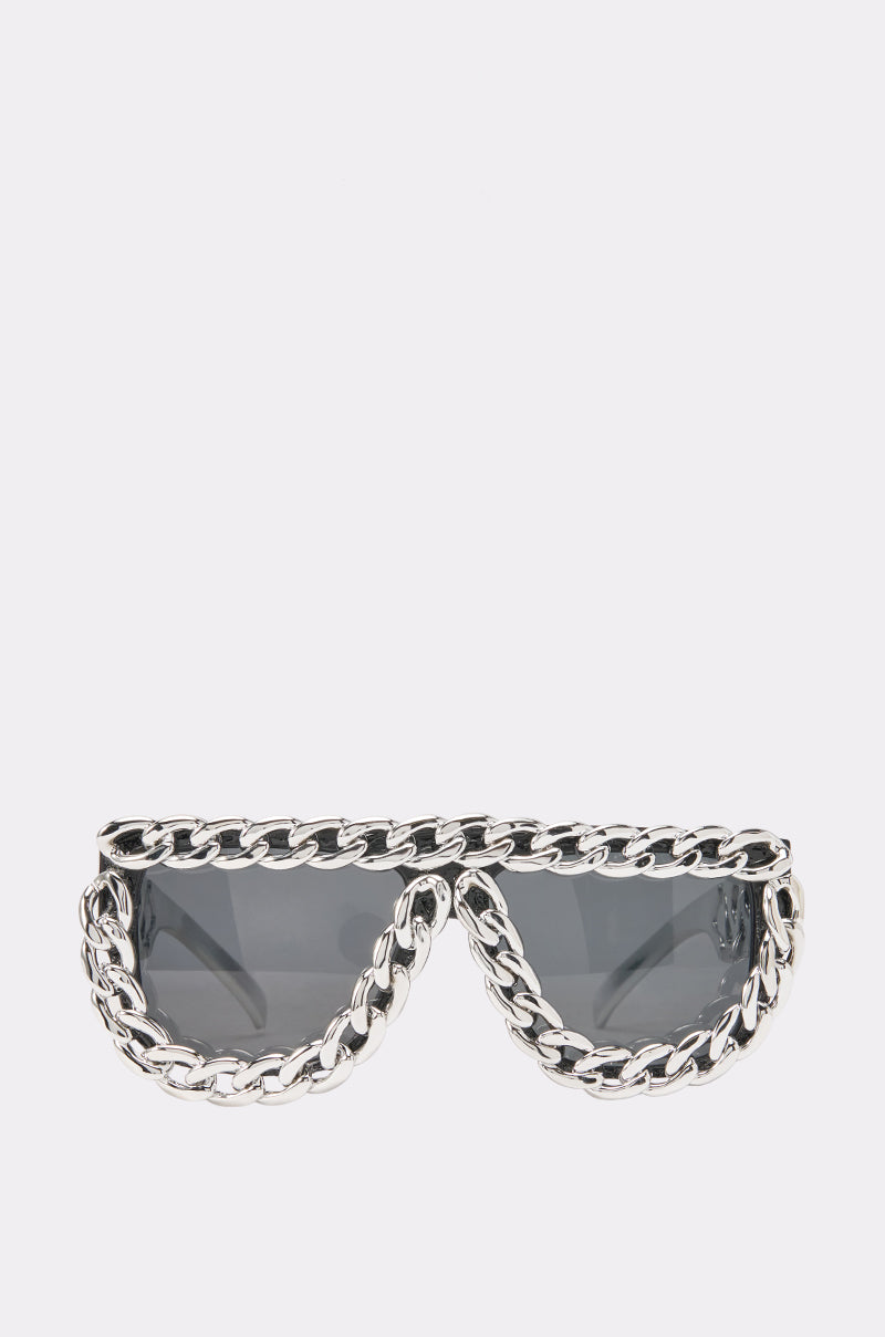 The LOCKED IN LUXURY SUNGLASSES feature dark lenses and are accented with a bold, thick silver chain outlining the entire frame for an edgy, streetwear-inspired look, shown against a plain white background.