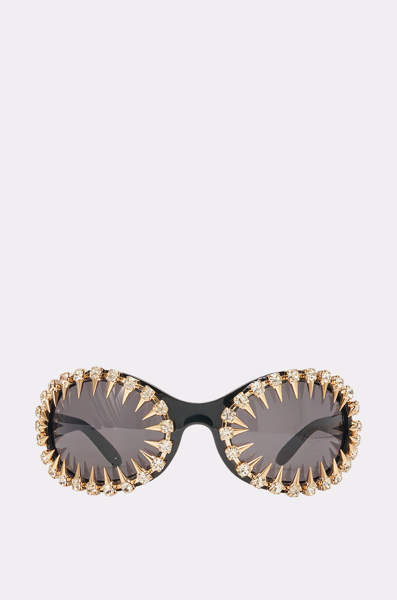 The GLAM SPIKE SUNGLASSES feature dark oval lenses and are adorned with gold spikes and rhinestones around the frames, creating a bold statement look with their dramatic, crown-like jagged border.
