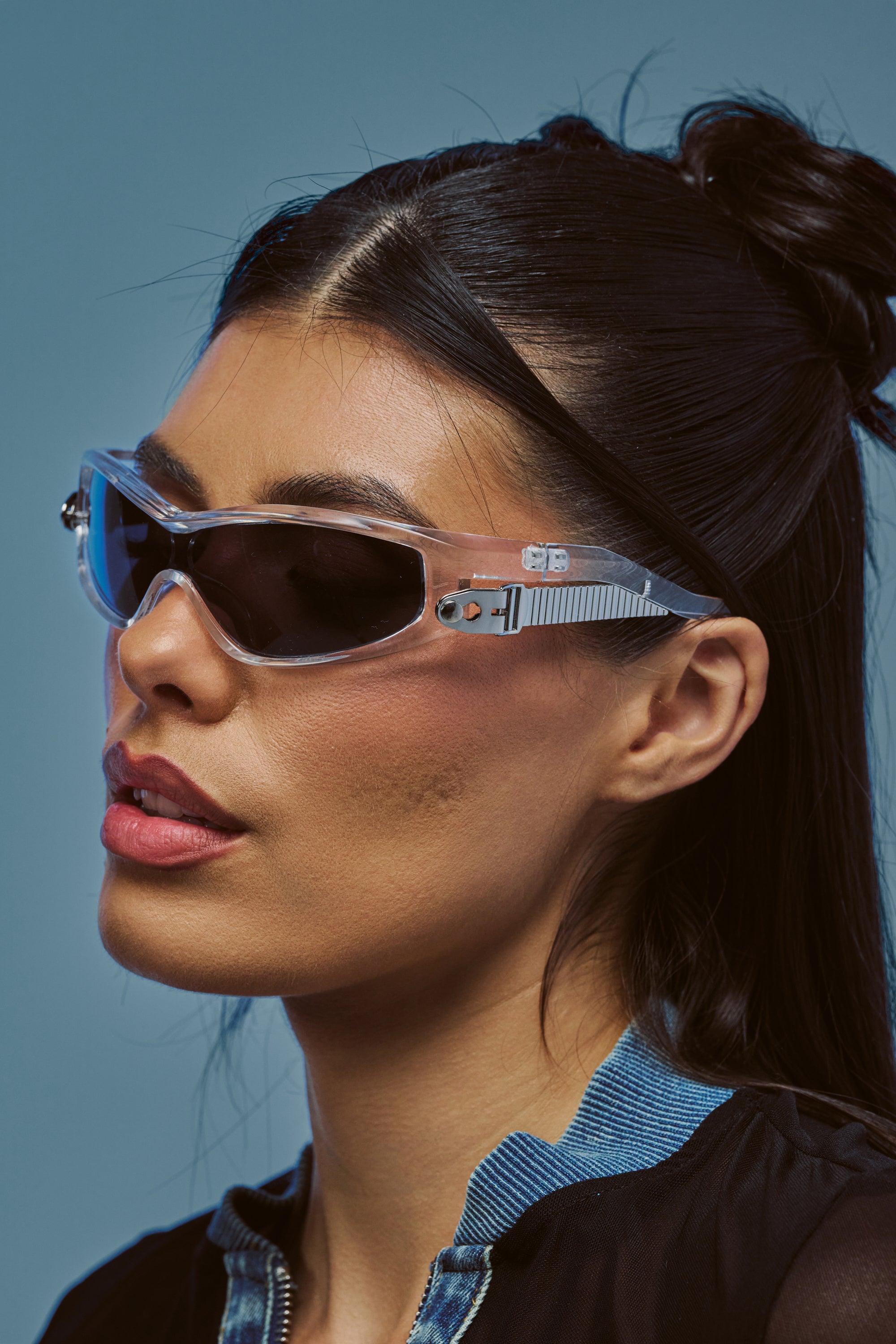 A woman with long, dark hair wears CLEAR CHAOS SUNGLASSES and a zip-up top, looking to the side against a blue background.