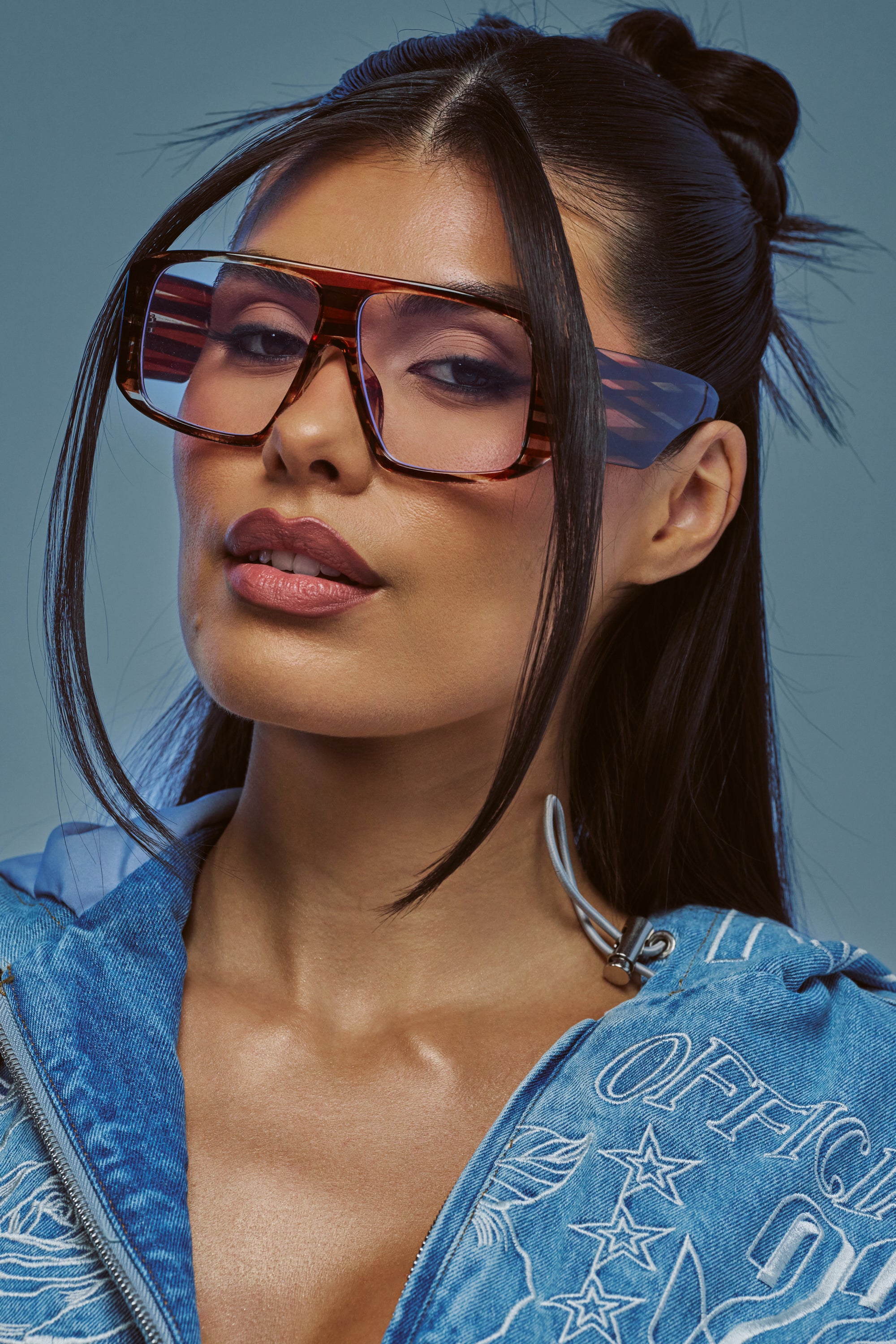A woman with long dark hair styled in half-up buns showcases the RETRO HEAT GLASSES—oversized, retro-inspired frames with brown stripes—while wearing a blue denim jacket with white embroidery and gazing confidently at the camera against a neutral background.