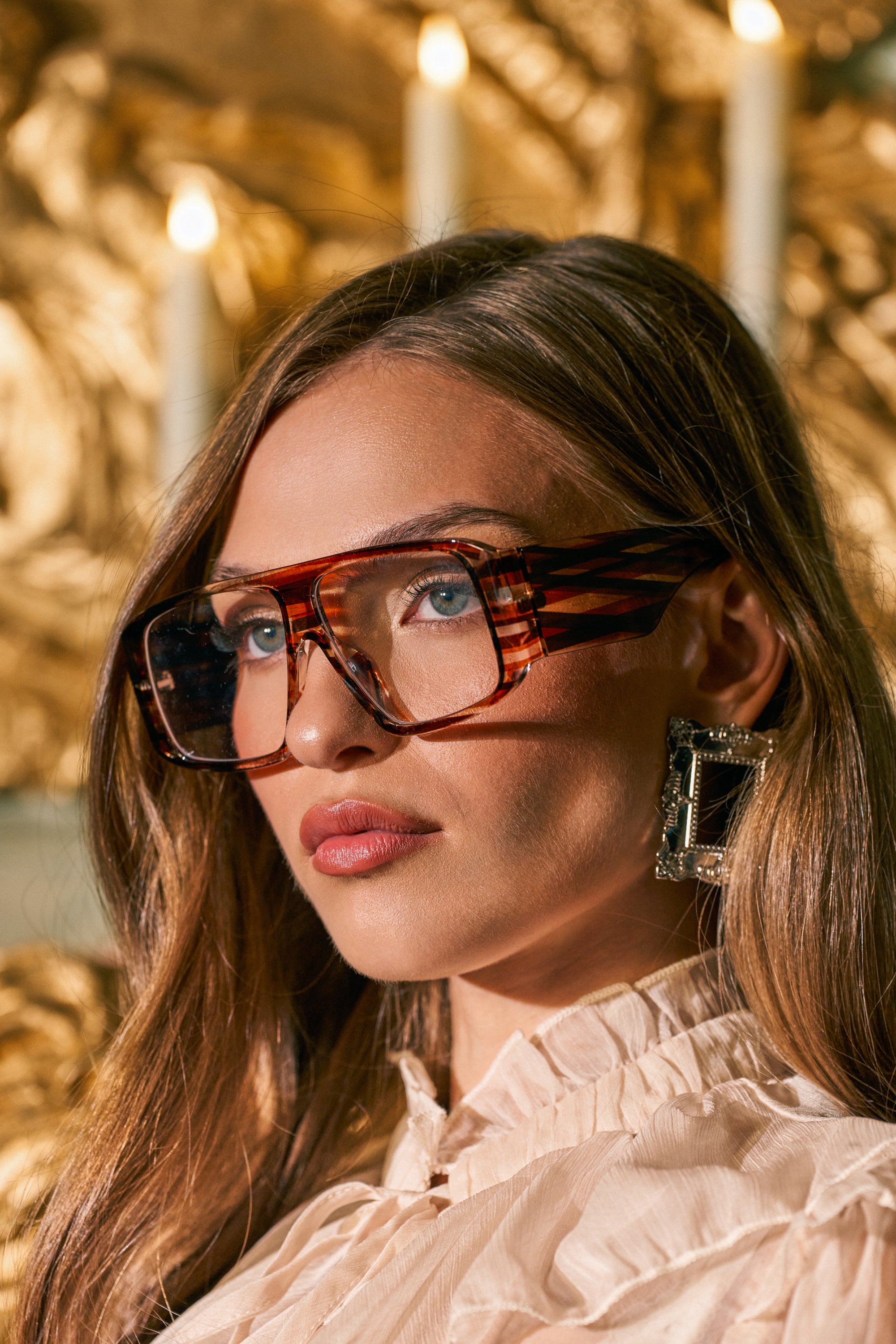 A woman with long hair wears the RETRO HEAT GLASSES—large, retro brown stripe frames—and square, silver statement earrings. She is dressed in a light ruffled top and poses in front of a gold, ornate background adorned with candles.