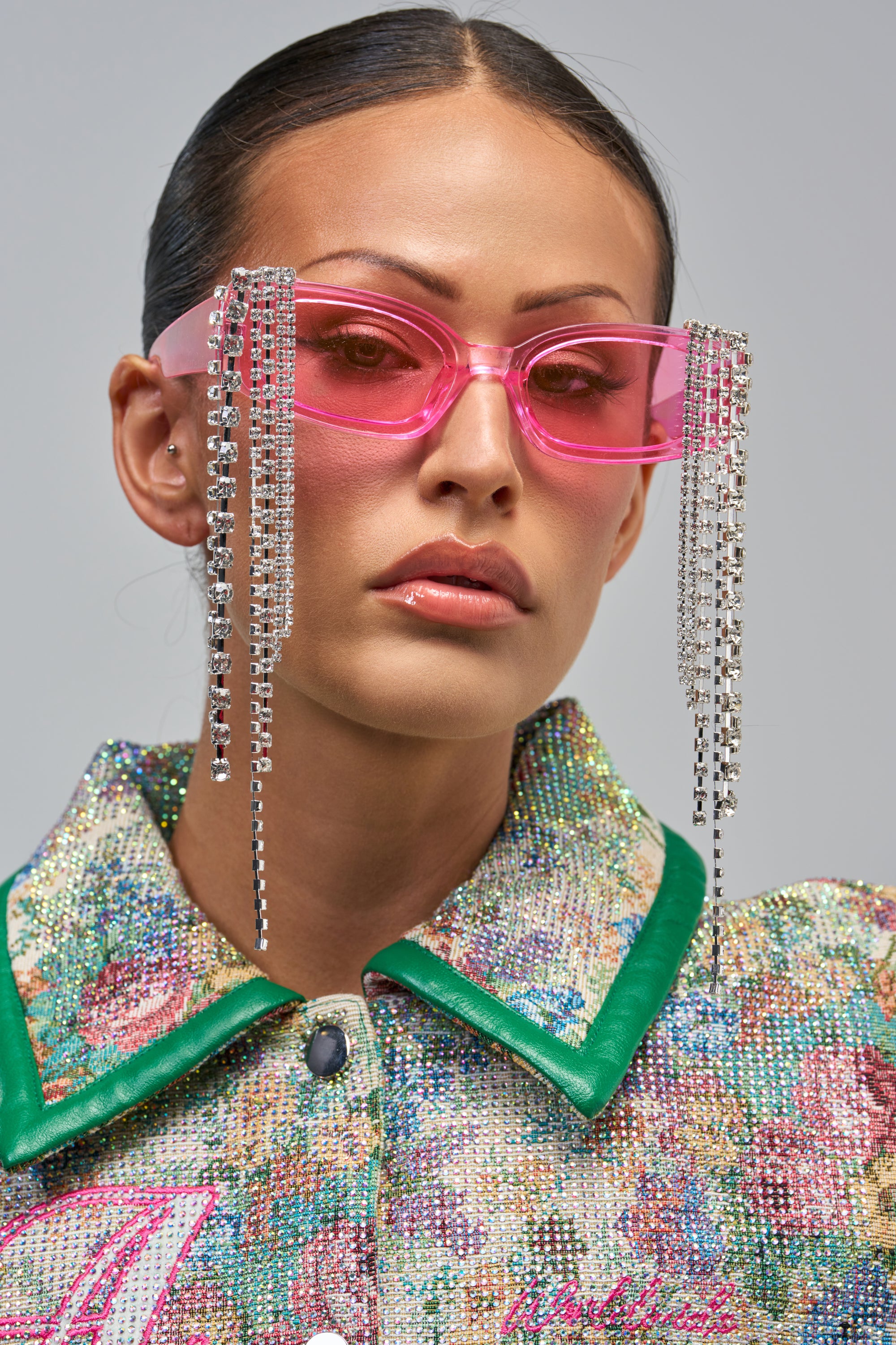 A woman wears BARBIE AFTER DARK SUNGLASSES with rhinestone fringe, long dangling chains, and a sparkly floral jacket with green trim. Her dark hair is pulled back and her lips are glossy.