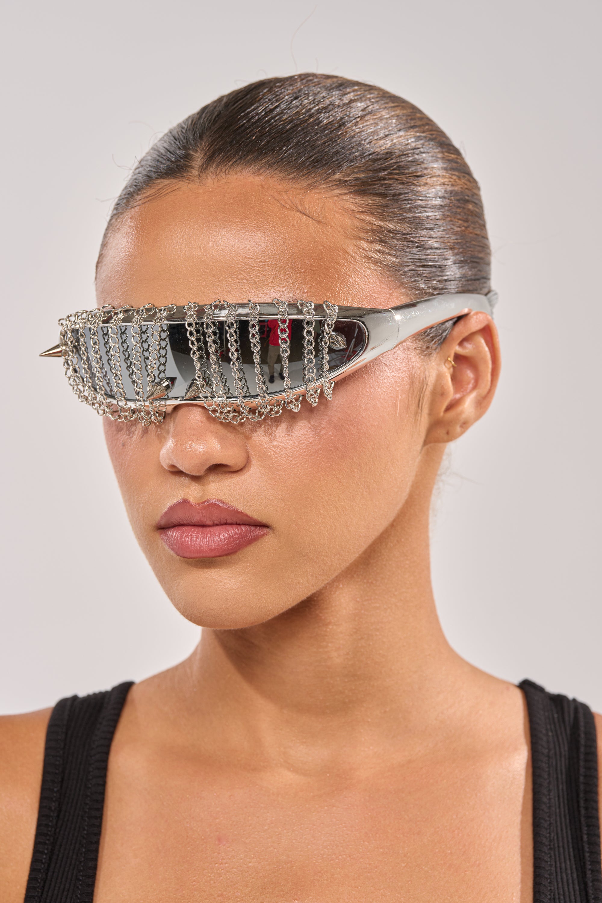 A woman with slicked-back hair wears CHAINED DOWN SUNGLASSES featuring a bold spike and chain design, paired with a black sleeveless top against a plain light backdrop. 1