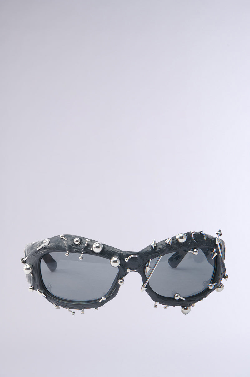 PIERCED VIBES SUNGLASSES in black, featuring multiple small silver-tone metallic details and round studs, displayed on a plain light grey background. 3