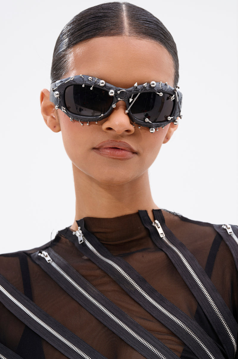 A woman with slicked-back hair wears PIERCED VIBES SUNGLASSES—black frames with silver-tone details—and a sheer black top with silver zippers, gazing confidently at the camera against a plain white background. 4