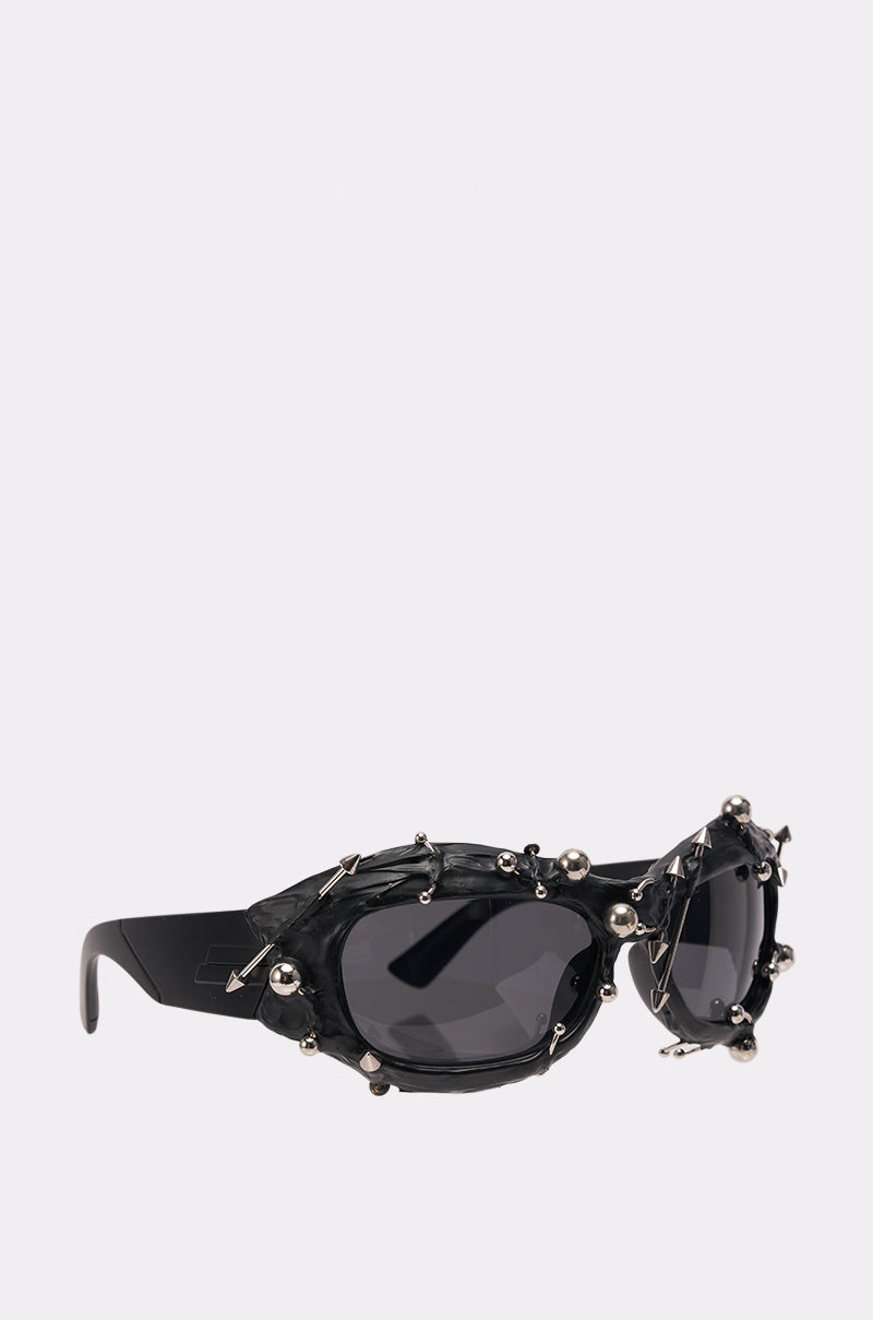 The PIERCED VIBES SUNGLASSES showcase oval lenses with small silver spikes and ball studs on black frames, adding a bold punk-rock edge. Displayed on a plain white background. 2