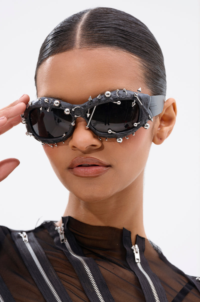 A woman with slicked-back hair wears PIERCED VIBES SUNGLASSES, featuring silver-tone metallic detailing, styled with a black mesh top with vertical zips. One hand is partially raised near her face in a gesture. 1
