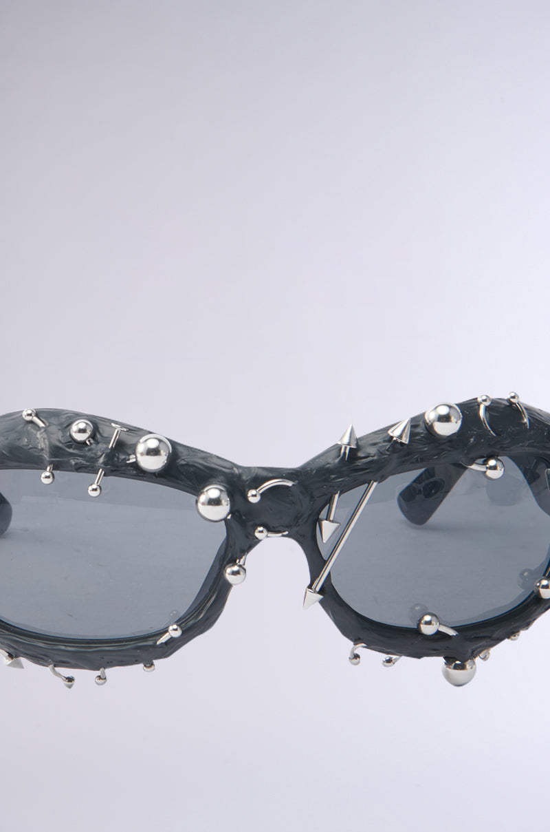 Close-up of PIERCED VIBES SUNGLASSES with tinted oval lenses, silver-tone metallic spikes, and round studs, displayed on a plain light gray background. 6