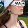 A woman poses outdoors in ICED OUT SUNGLASSES featuring a wavy black pattern, a white beanie, metallic hand-shaped earrings, and a black strappy top with buildings and greenery behind her.