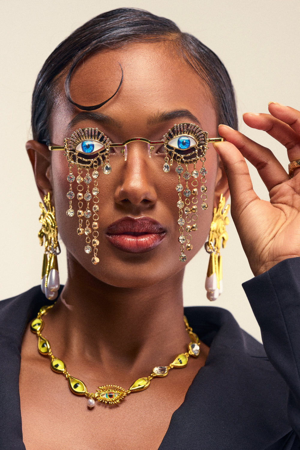 A woman showcases the EYES ON ME SUNGLASS—gold avant-garde eyewear embellished with realistic blue eyes and dangling jewels—completing her statement look with gold earrings and a gold necklace. She holds the distinctive sunglasses in one hand, pairing them with a dark blazer against a neutral background.