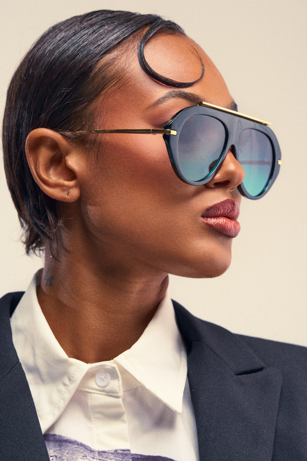 A person with short slicked hair wears the LUXE AIR SUNGLASSES IN BLACK BLUE—featuring oversized aviator frames and black-and-blue reflective lenses—paired with a black blazer and white collared shirt, looking to the side against a plain background.