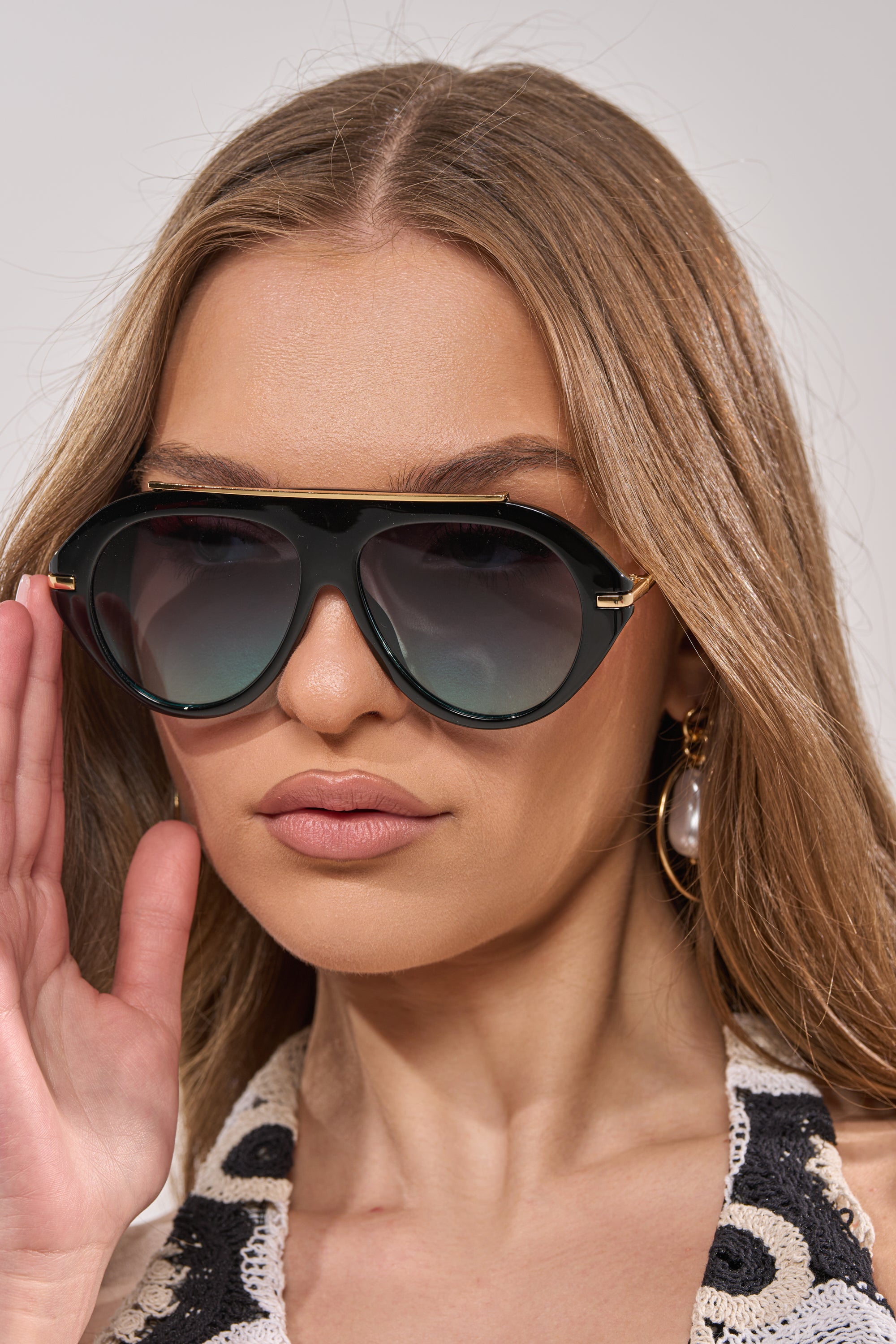 A woman with long light brown hair wears LUXE AIR SUNGLASSES IN BLACK BLUE, a patterned top, and a pearl earring. She confidently looks forward with one hand raised near her face. 2