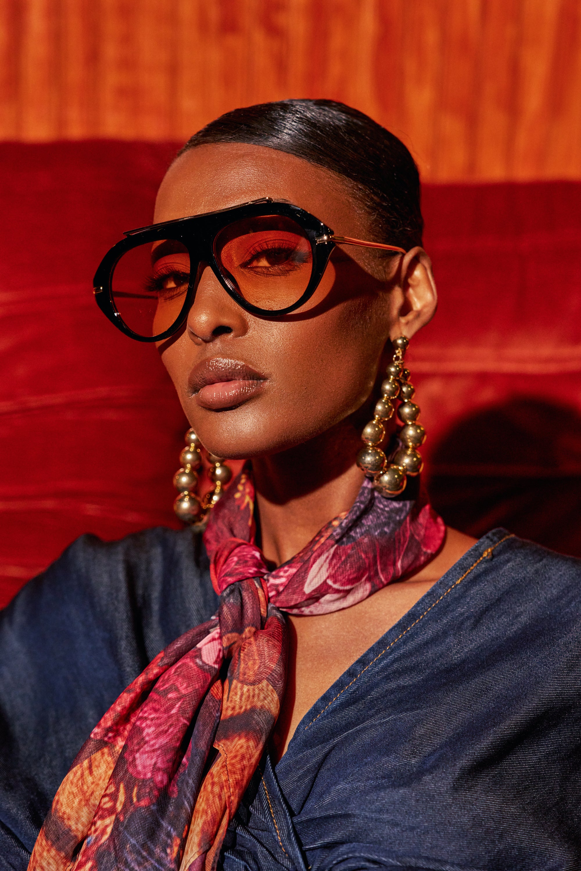 A person wearing LUXE AIR SUNGLASSES IN BLACK ORANGE, large gold spherical earrings, and a patterned scarf.