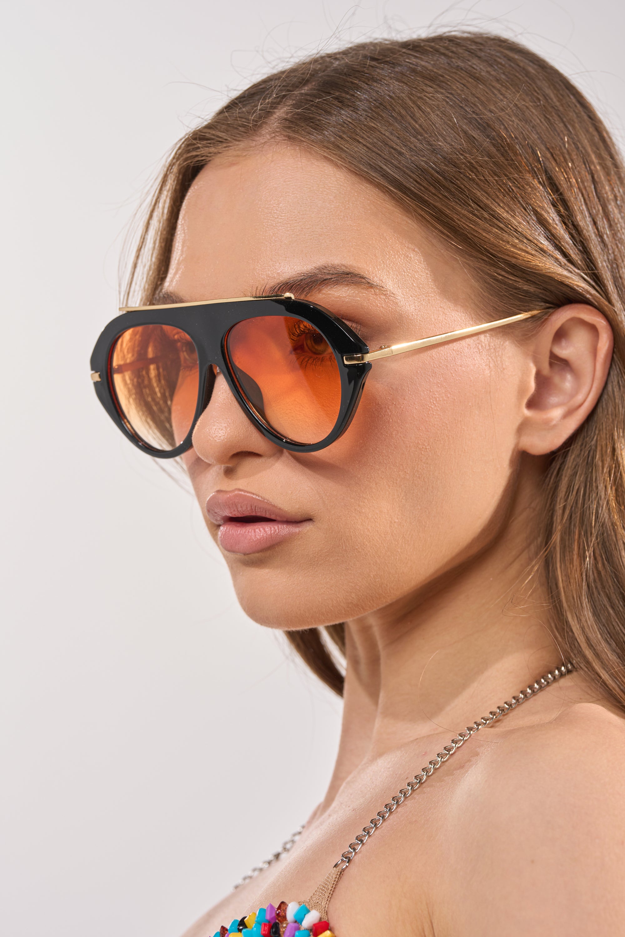 A woman with long brown hair and a neutral expression wears LUXE AIR SUNGLASSES IN BLACK ORANGE, styled with oversized aviator frames. She is dressed in a colorful beaded top featuring a silver chain strap. 2